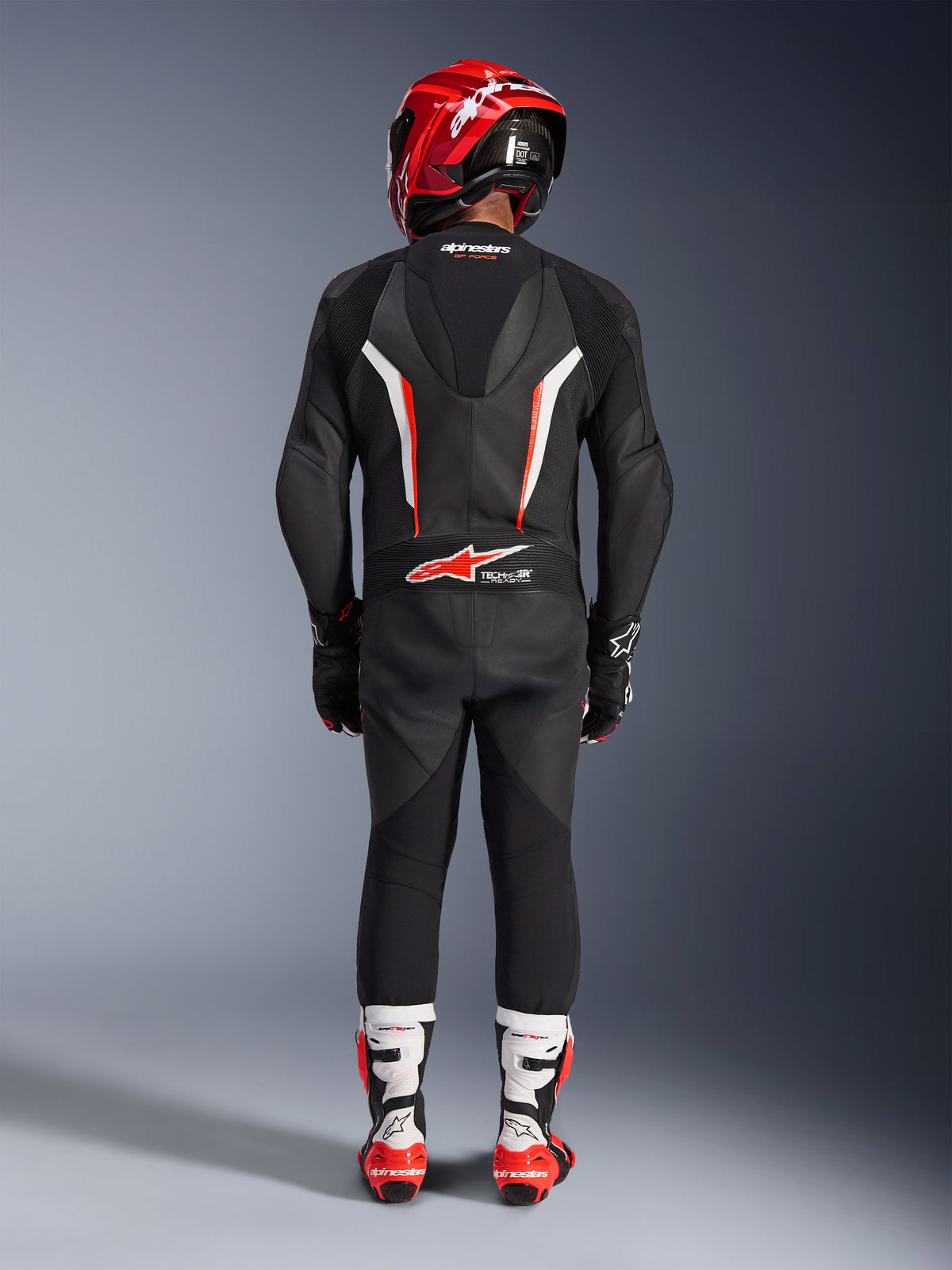 Fahrer in der Rückansicht in der Alpinestars GP Force V2 1-Teiler Lederkombi in Schwarz Weiß Rot Fluo, mit aerodynamischem Geschwindigkeitsbuckel und Tech-Air Ready-Branding, kombiniert mit einem roten Rennhelm und weiß-roten Stiefeln, professionelles Design für die Rennstrecke.