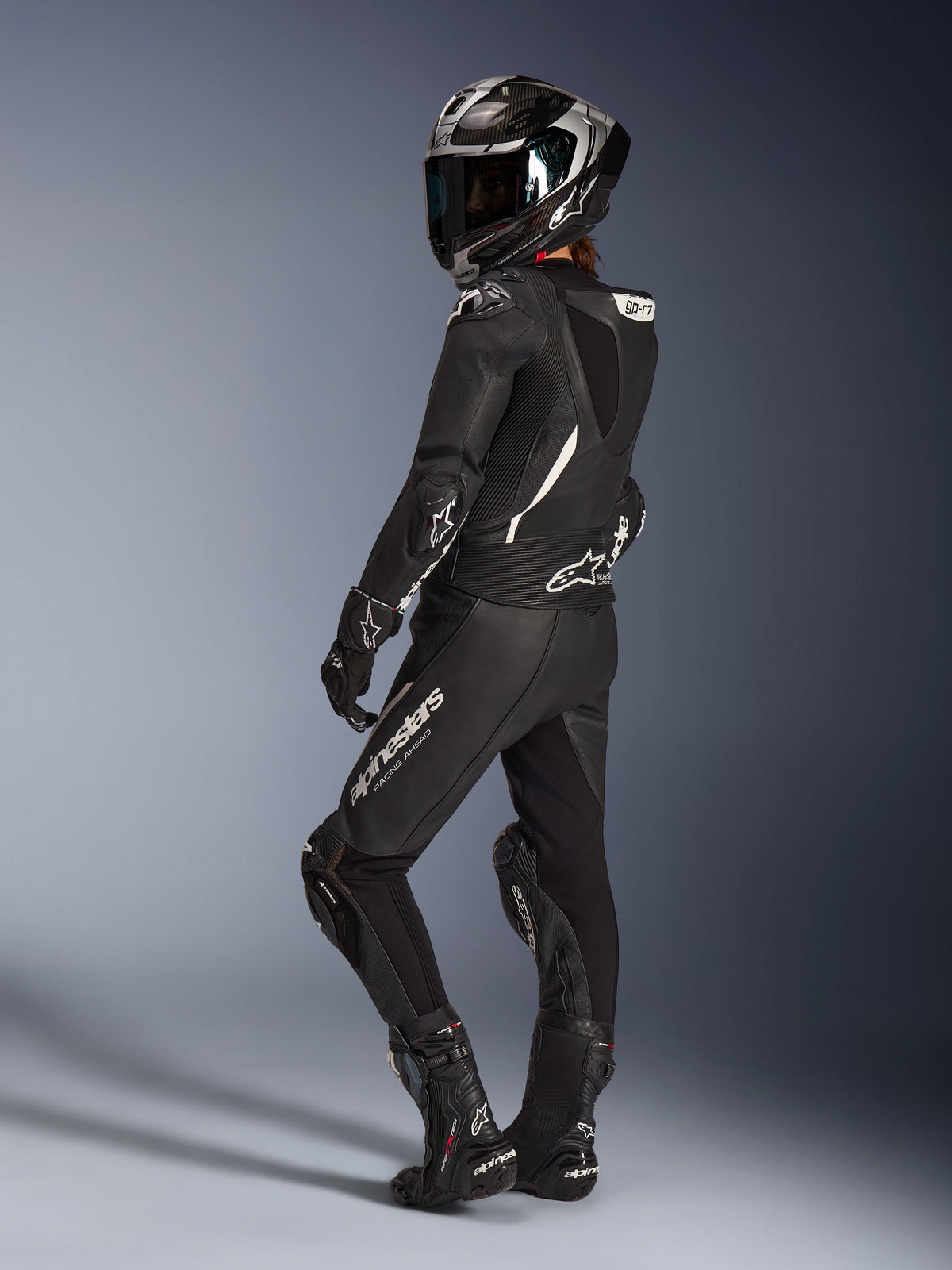 Fahrerin im schwarzen Alpinestars Stella GP-R7 2-Teiler Lederanzug, Integral-Rennhelm mit Carbon-Finish, passende Handschuhe und Stiefel, Rückansicht mit aerodynamischem Höcker und weißen Alpinestars-Logos, professionelle Rennstrecken- und Sport-Fahrausrüstung