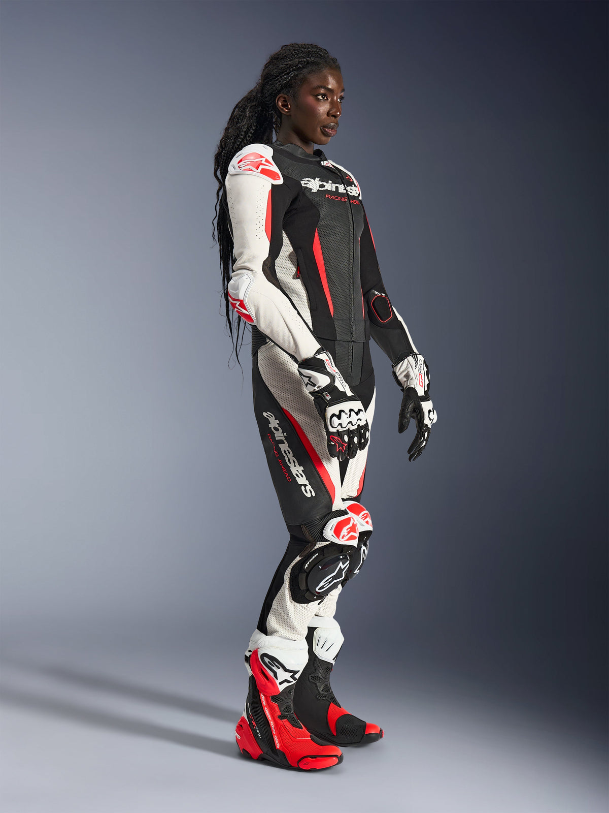 Motorradfahrerin im Alpinestars Stella GP-R7 2-Teiler-Lederanzug in Schwarz, Weiß und Red Fluo, kombiniert mit passenden Rennhandschuhen und Stiefeln, mit perforierten Lederbahnen und Protektoren an Schultern, Ellbogen und Knien, stehend in einer Studioumgebung.