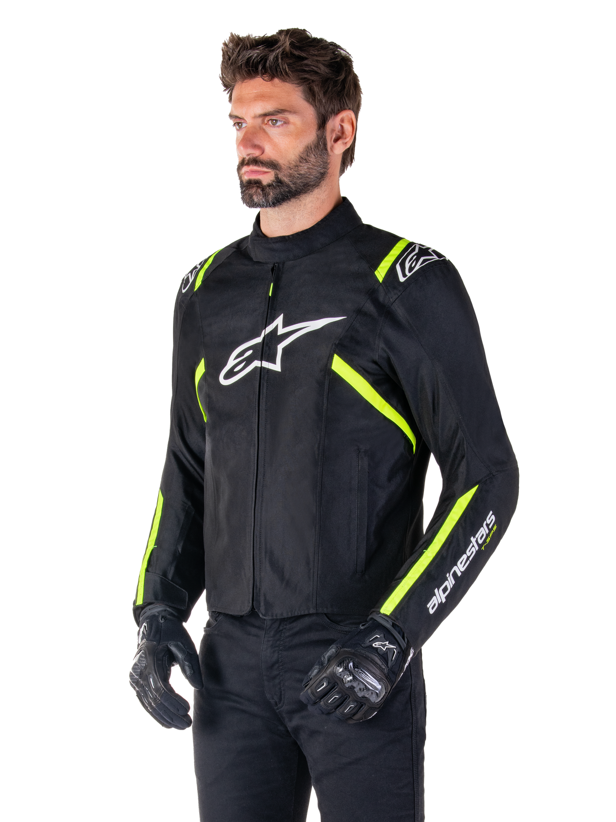 Männliches Model in der Alpinestars T-SPS V2 Waterproof Jacke, Schwarz mit weißen und gelben Fluo-Akzenten, mit einem weißen Astars-Logo auf der Brust und fluoreszierenden gelben Streifen an den Schultern und Ärmeln, kombiniert mit einer schwarzen Hose und Motorradhandschuhen, sportlich geschnittene Textil-Motorradjacke.