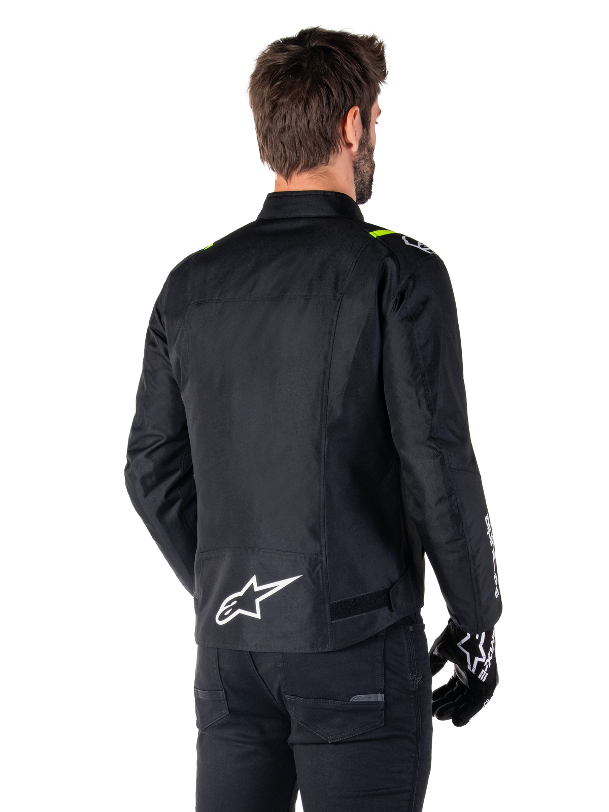 Alpinestars T-SPS V2 Waterproof Jacke, Schwarz Weiß Gelb Fluo, Rückansicht eines männlichen Models, das die Textil-Motorradjacke trägt, mit einem weißen Logo am unteren Rücken und gelben Fluo-Akzenten an den Schultern, kombiniert mit schwarzer Hose und Handschuhen.
