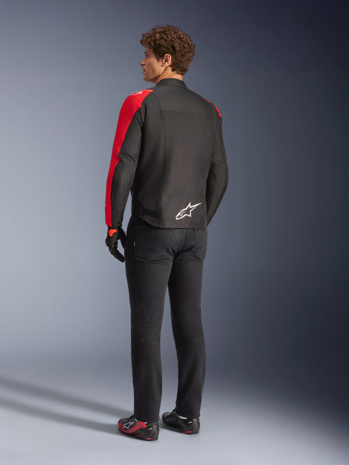 Alpinestars T-SPS V2 Waterproof Jacket in Black and Red Fluo, rear view on model, worn with black motorcycle pants and riding shoes, featuring a white Alpinestars logo on the lower back, sport-cut textile jacket design.