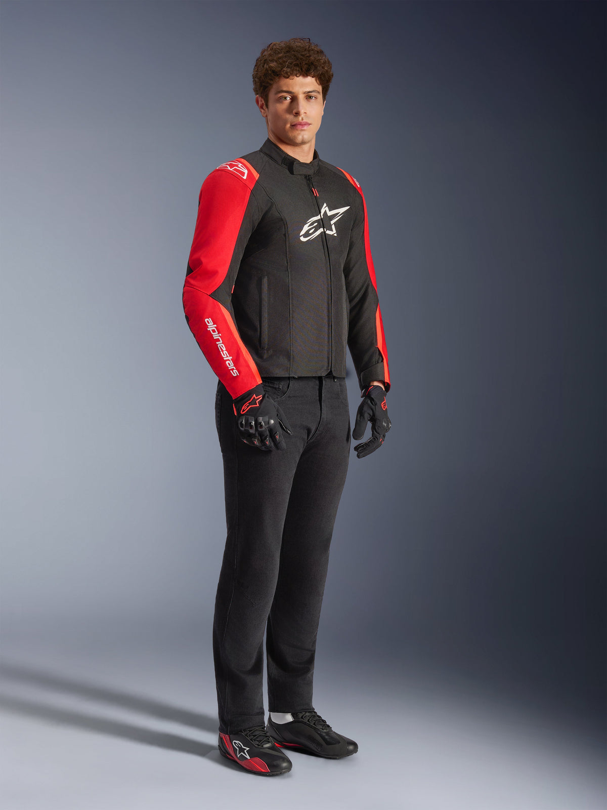 Male model wearing Alpinestars T-SPS V2 Waterproof Jacket in Black and Red Fluo, featuring vibrant red sleeves with white branding and a large white logo on the chest, paired with black riding pants, motorcycle gloves, and low-profile riding shoes, full-length studio view.