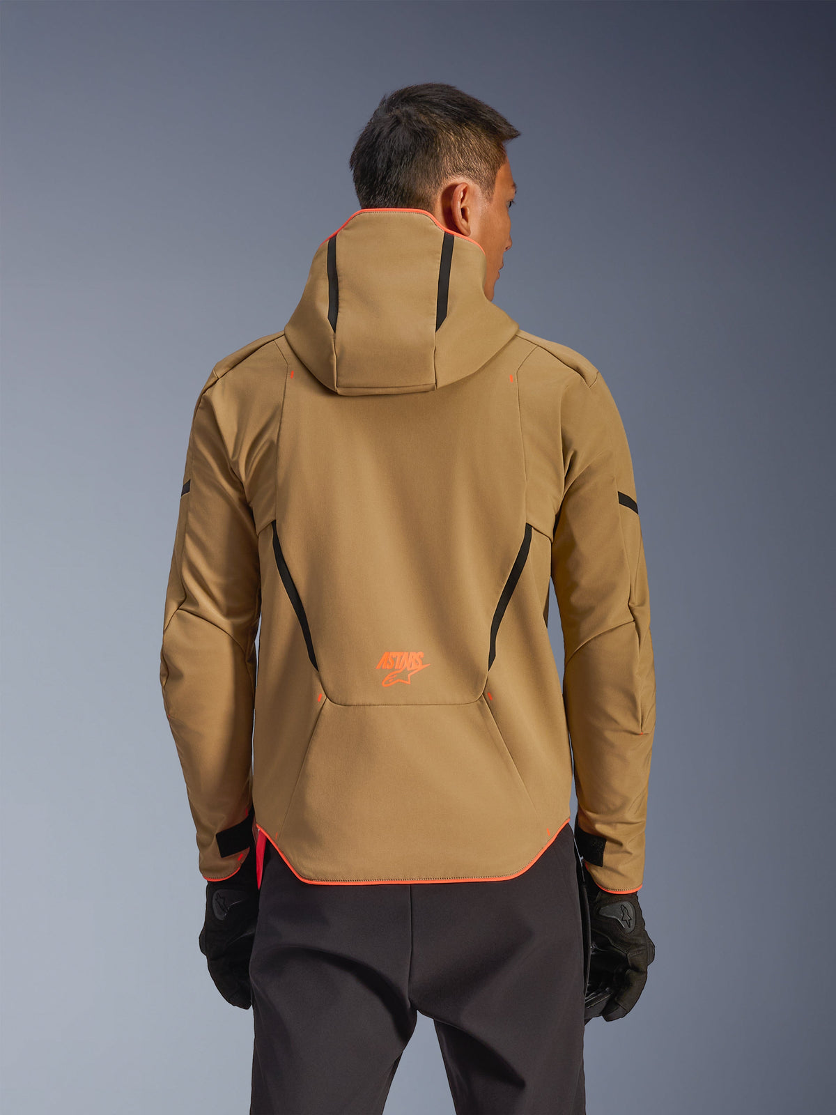 Alpinestars Aeroshell WP Jacke, wasserdichte Jacke, Dark Khaki und Orange Fluo, Rückansicht zeigt Hoodie-Design mit orangefarbenem Besatz an Kapuze und Saum, orangefarbenes Logo am unteren Rücken, getragen mit schwarzen Handschuhen, Crossover-Sport-Softshell-Motorradausrüstung