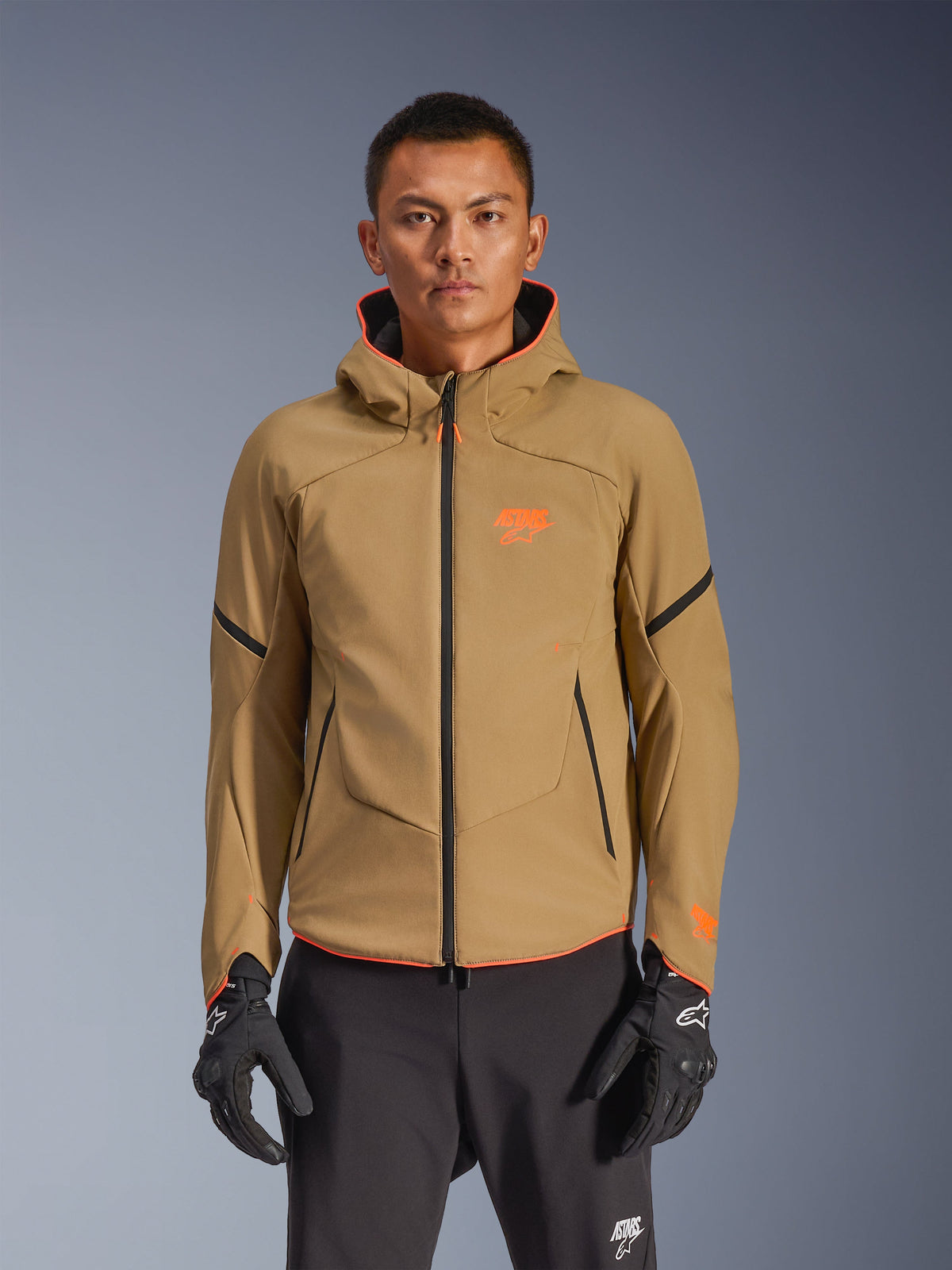 Alpinestars Aeroshell Waterproof Jacke, wasserdichte Motorradjacke mit Kapuze in Dark Khaki mit orangefarbenen Logos und schwarzen Reißverschlüssen, schwarze Akzentlinien an den Ärmeln, abgebildet mit schwarzen Handschuhen und Hosen