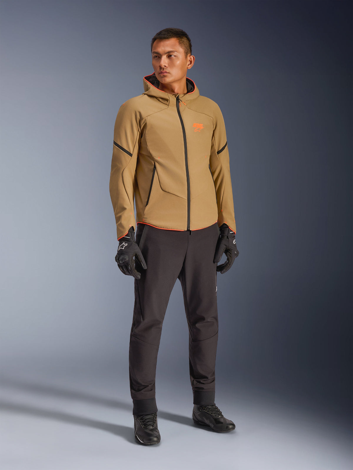 Alpinestars Aeroshell wasserdichte Jacke in Dark Khaki, Schwarz und Orange Fluo, getragen von einem Modell mit passender Ausrüstung, Kapuzen-Softshell-Design, wasserdichte Reißverschlüsse, leuchtend orangefarbene Akzente und Branding