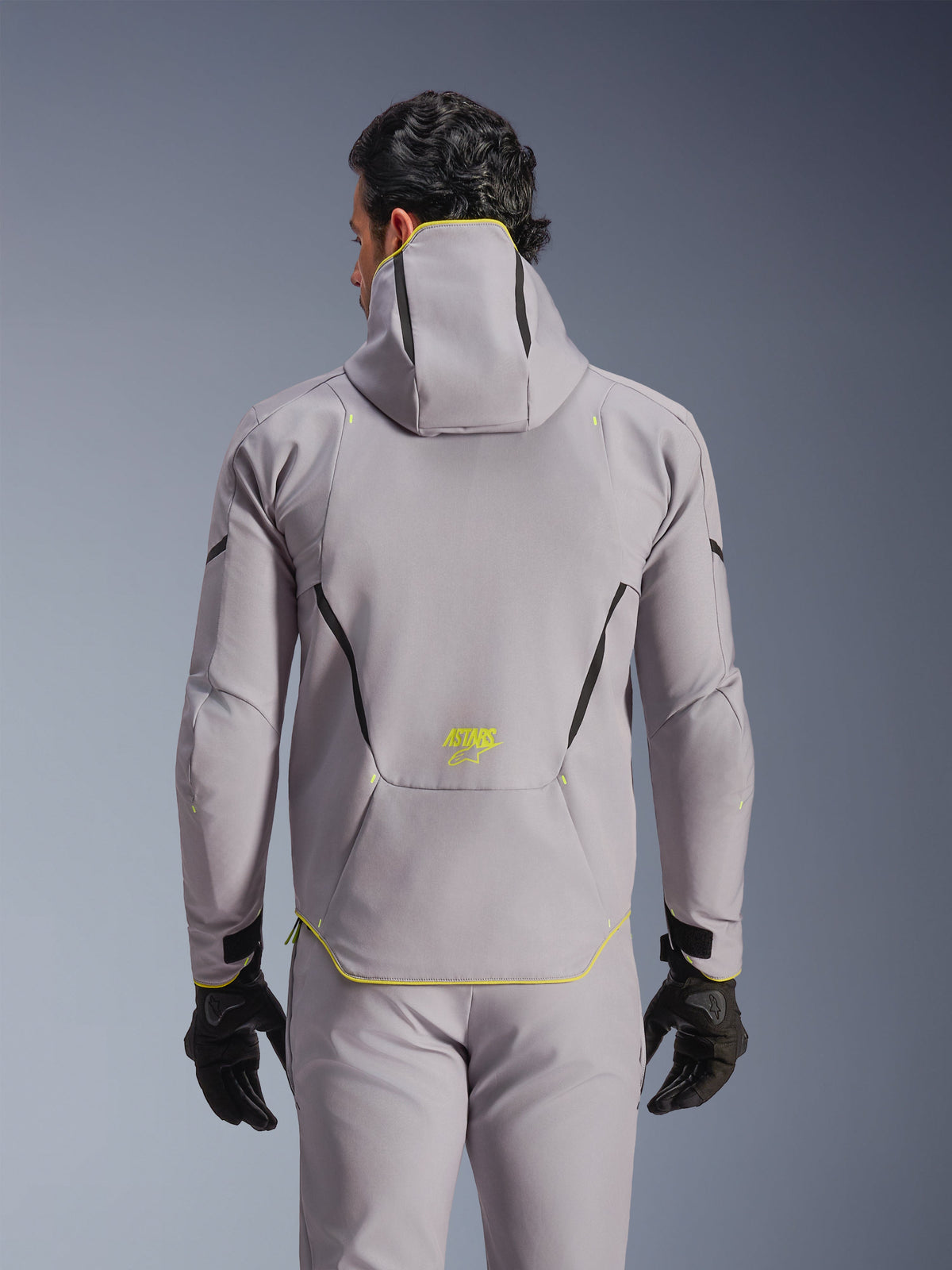 Alpinestars Aeroshell WP Jacke, Mittelgrau mit neongelben Akzenten, Rückansicht mit integrierter Kapuze und schwarzen Kontrastlinien, gelbes ASTARS-Logo am unteren Rücken, getragen mit passender grauer Hose und schwarzen Handschuhen, Sport-Softshell-Motorradjacke