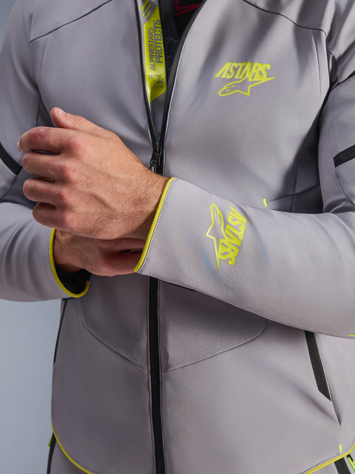 Alpinestars Aeroshell WP Jacke, Mittelgrau mit gelben Fluo-Akzenten, Nahaufnahme von Brust und Ärmelbündchen mit fluoreszierenden gelben Astars-Logos, Detail der Innenklappe mit Tech-Air Ready-Branding, moderne Sport-Softshell-Motorradjacke