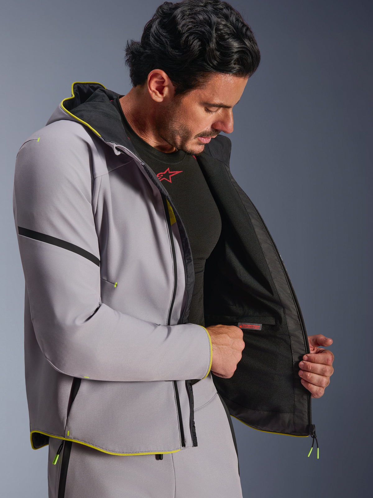 Alpinestars Aeroshell WP Jacke in Mittelgrau und Fluorgelb, getragen von einem männlichen Modell, zeigt das interne wasserdichte Fach und das schwarze Innenfutter, Sport-Softshell-Motorrad-Hoodie mit fluoreszierend gelben Details und moderner sportlicher Passform