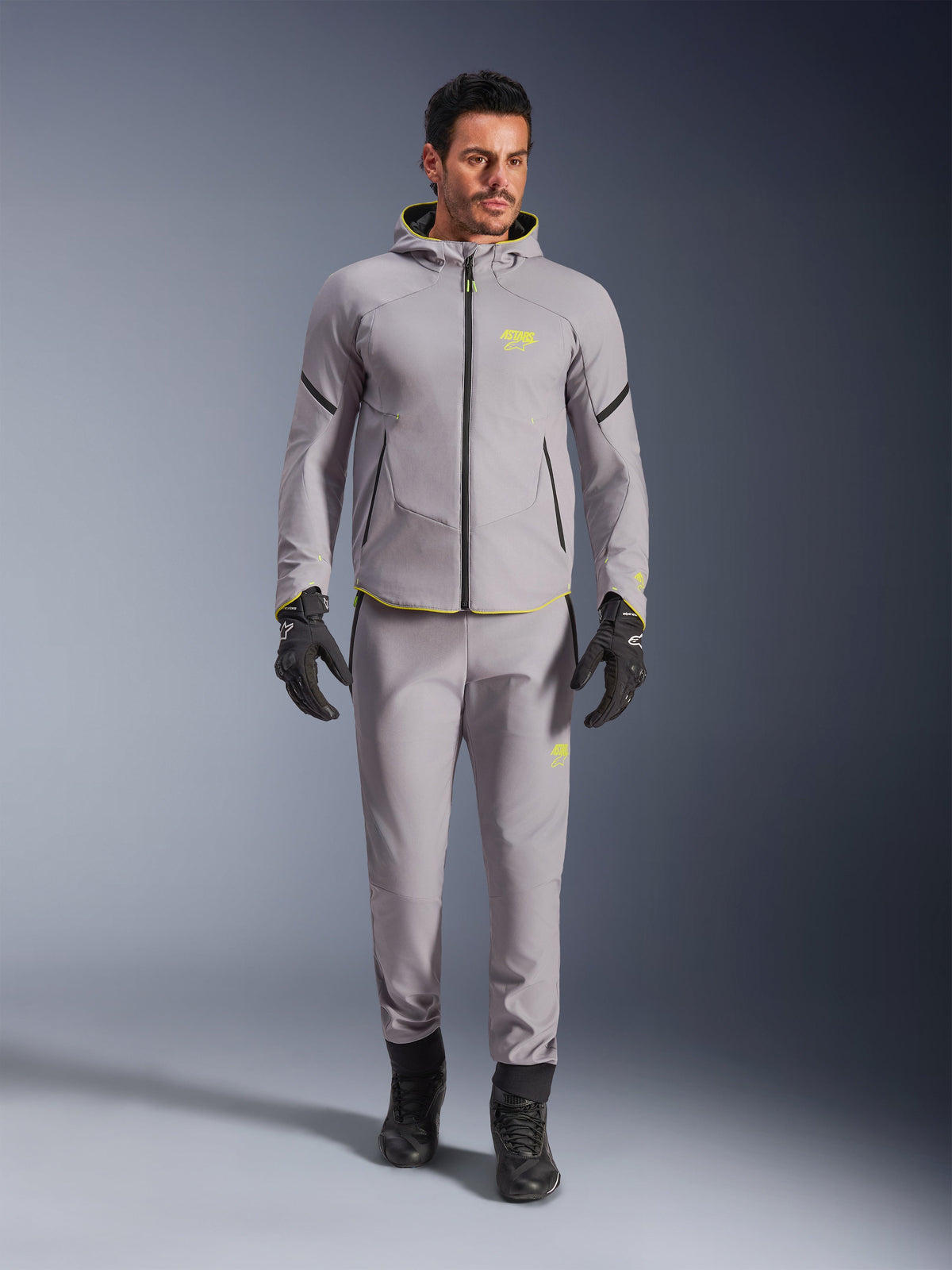 Alpinestars Aeroshell Waterproof Jacke, Mittelgrau Gelb Fluo, getragen mit passender grauer Hose, schwarzen Handschuhen und Stiefeln, modernes sportlich inspiriertes Hoodie-Design mit leuchtend gelben Akzenten und athletischer Passform