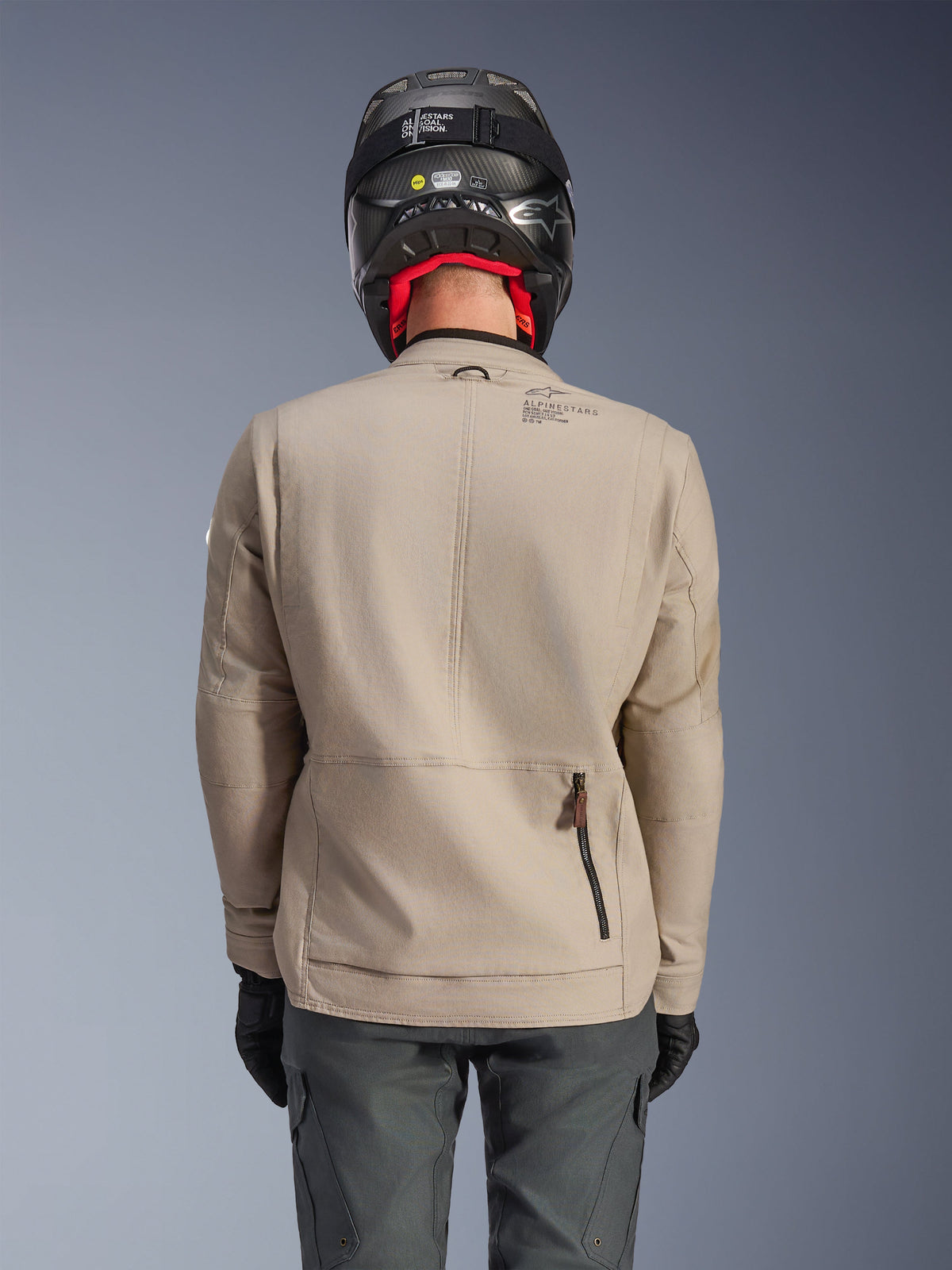 Alpinestars Borrego Drystar® Jacke, Adventure-Motorradjacke, Warm Gray, Rückansicht mit Reißverschluss am Rückenfach und Alpinestars-Branding, getragen mit schwarzem Helm und anthrazitfarbener Motorradhose