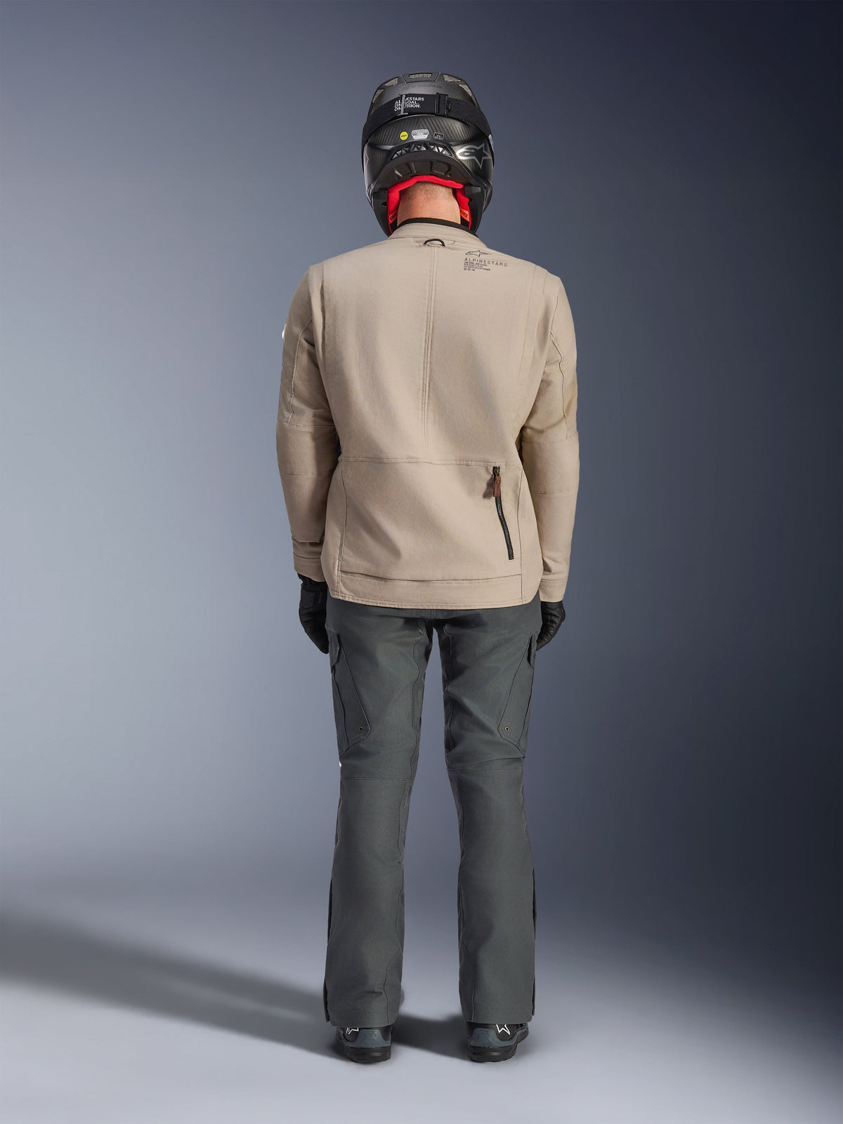 Alpinestars Borrego Drystar® Jacke, Adventure-Motorradjacke in Warm Gray, Rückansicht mit gewachstem Canvas-Look, Dehnungseinsätzen an den Schultern und einer Reißverschlusstasche am unteren Rücken, getragen mit dunkelgrauer Motorradhose und schwarzem Helm.