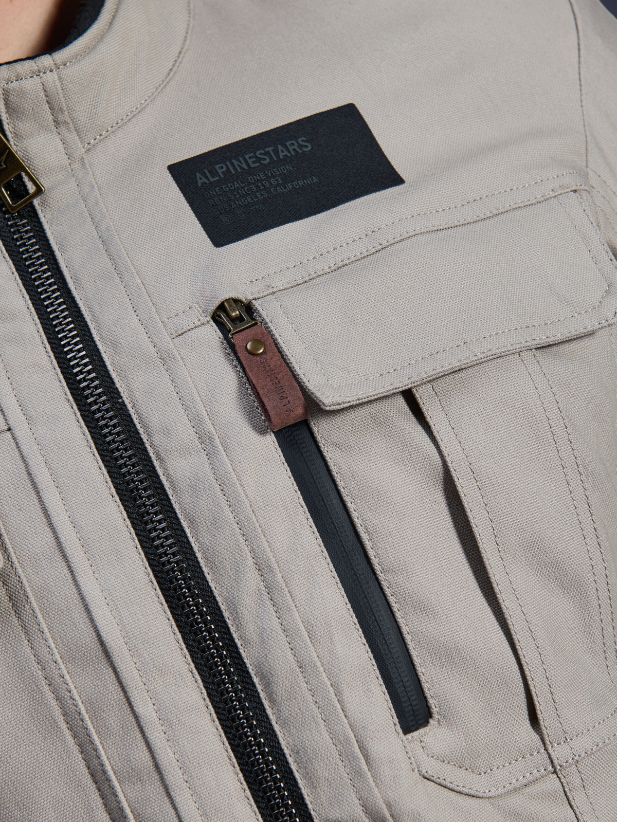 Alpinestars Borrego Drystar® Jacke, Warm Gray, Nahaufnahme des Brustbereichs mit einem schwarzen Alpinestars-Markenaufnäher, Brusttasche mit Klappe und einem vertikalen Reißverschluss mit brauner Lederlasche, robuste graue Textil-Canvas-Konstruktion.