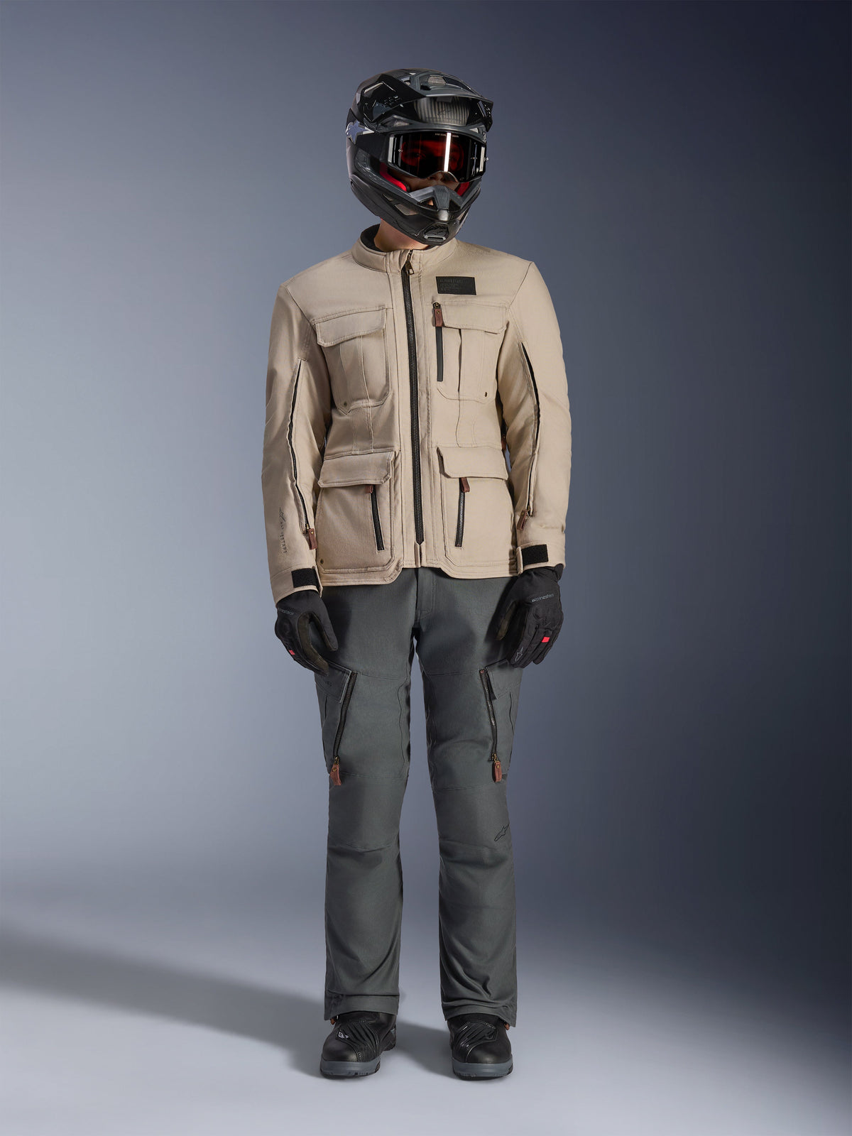 Alpinestars Borrego Drystar® Jacke in Warm Gray, Adventure-Motorradjacke mit vier Cargo-Taschen auf der Vorderseite, kombiniert mit dunkelgrauer Fahrhose, schwarzem Offroad-Helm und schwarzen Handschuhen, im robusten gewachsten Canvas-Look