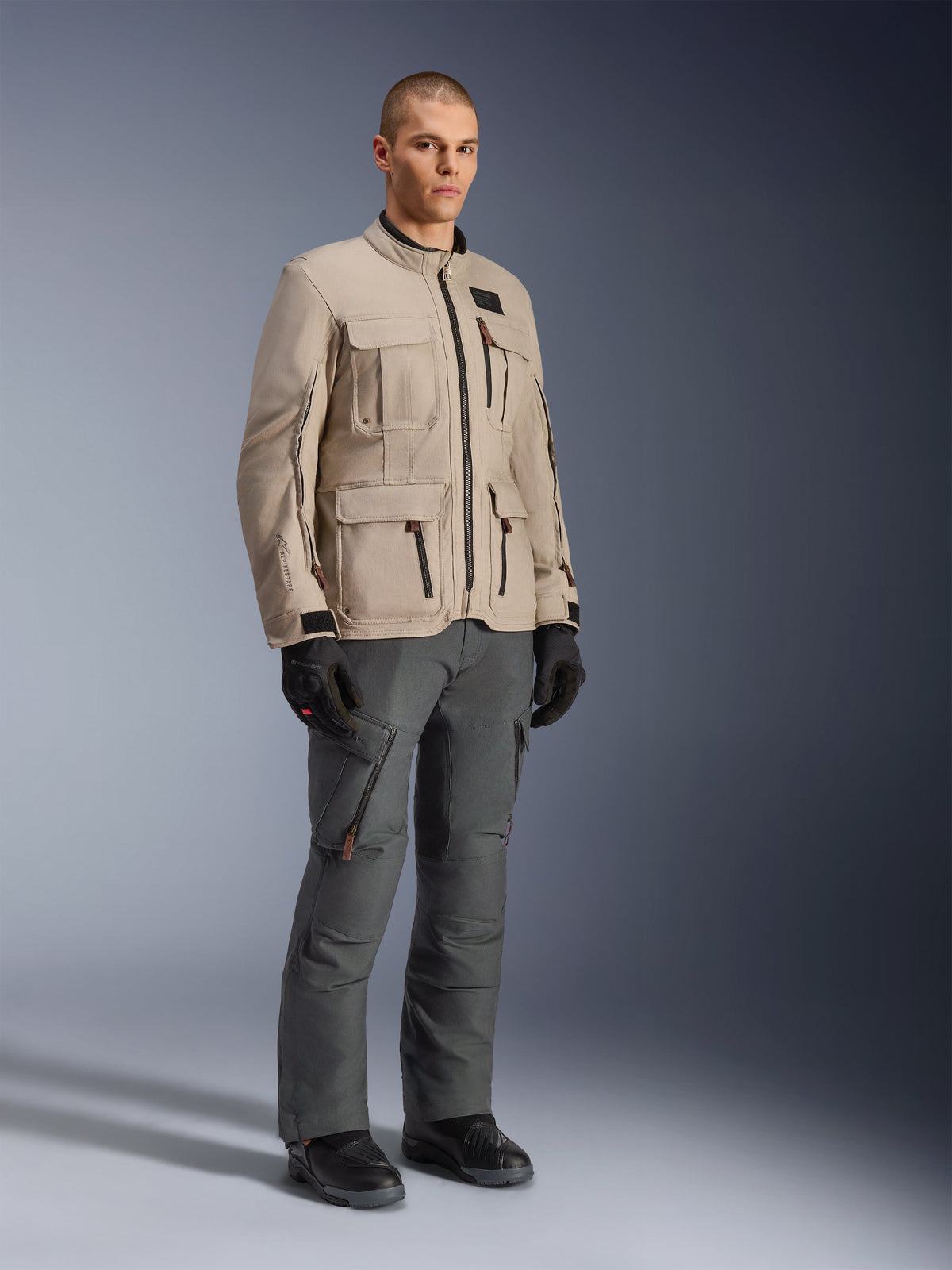 Alpinestars Borrego Drystar® Jacke in Warm Gray, Adventure-Motorradjacke mit gewachster Canvas-Optik und mehreren Funktionstaschen, getragen von einem Model mit dunkelgrauer Motorradhose, schwarzen Handschuhen und schwarzen Stiefeln, Ganzkörper-Studioporträt
