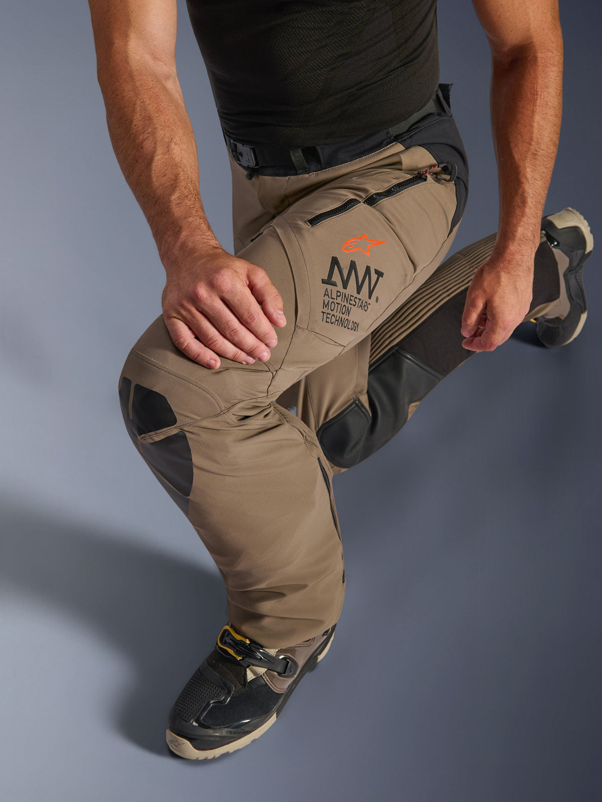 Alpinestars AMT-8 Stretch Drystar® XF Hose in Walnut Black, mit Alpinestars Motion Technology (AMT) Branding am Oberschenkel, langen Belüftungsöffnungen mit Reißverschluss und strapazierfähigen Kniepaneelen aus Leder, an einem knienden Fahrer zur Demonstration der Flexibilität.