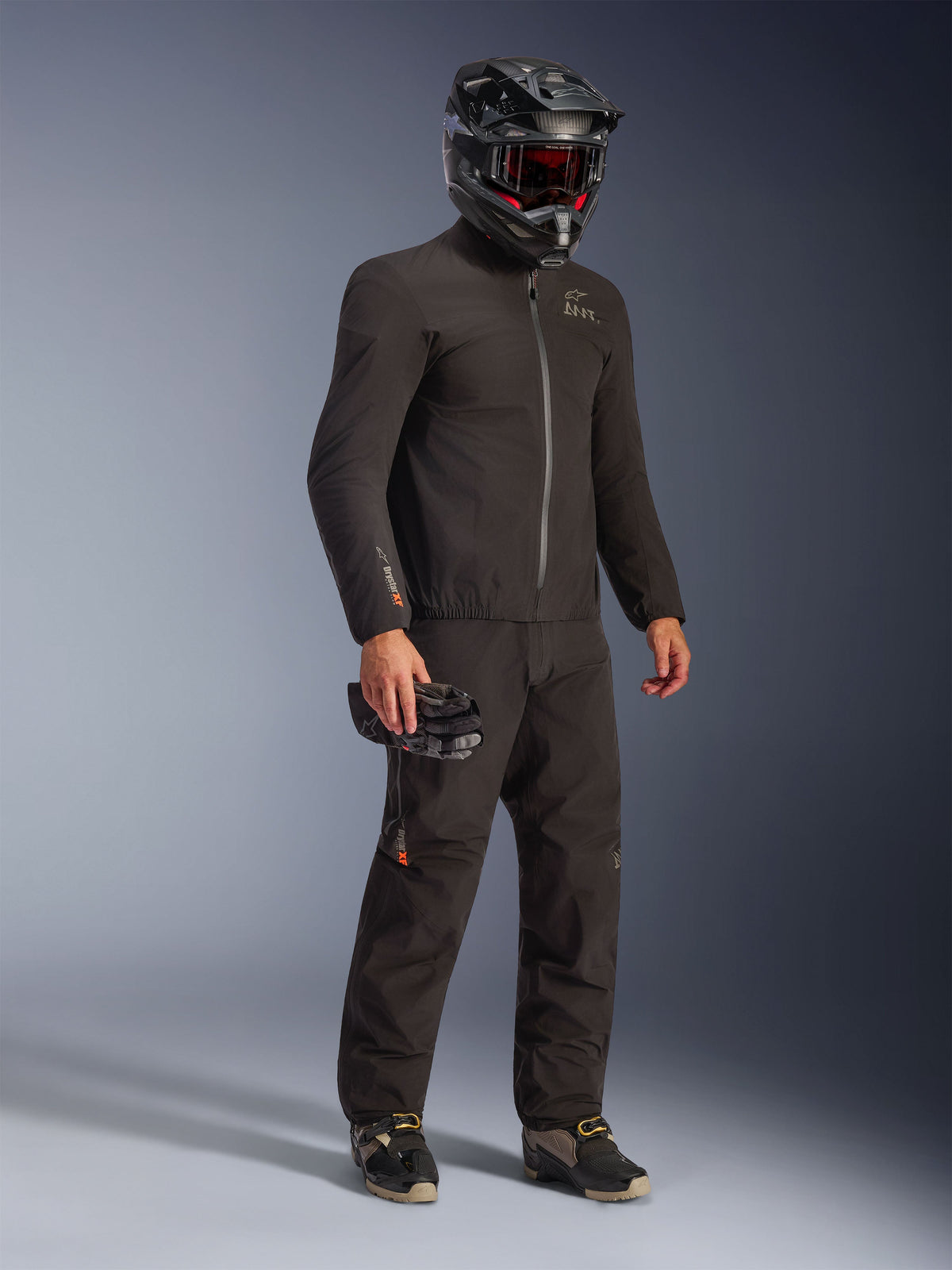 Alpinestars AMT-8 Stretch Drystar® XF Jacke in Schwarz, getragen als Teil einer kompletten Fahrerausrüstung mit passender Hose und schwarzem Helm, elegantes Textildesign mit orangefarbenem Drystar XF-Branding am Ärmel, ideal für Adventure-Touring.