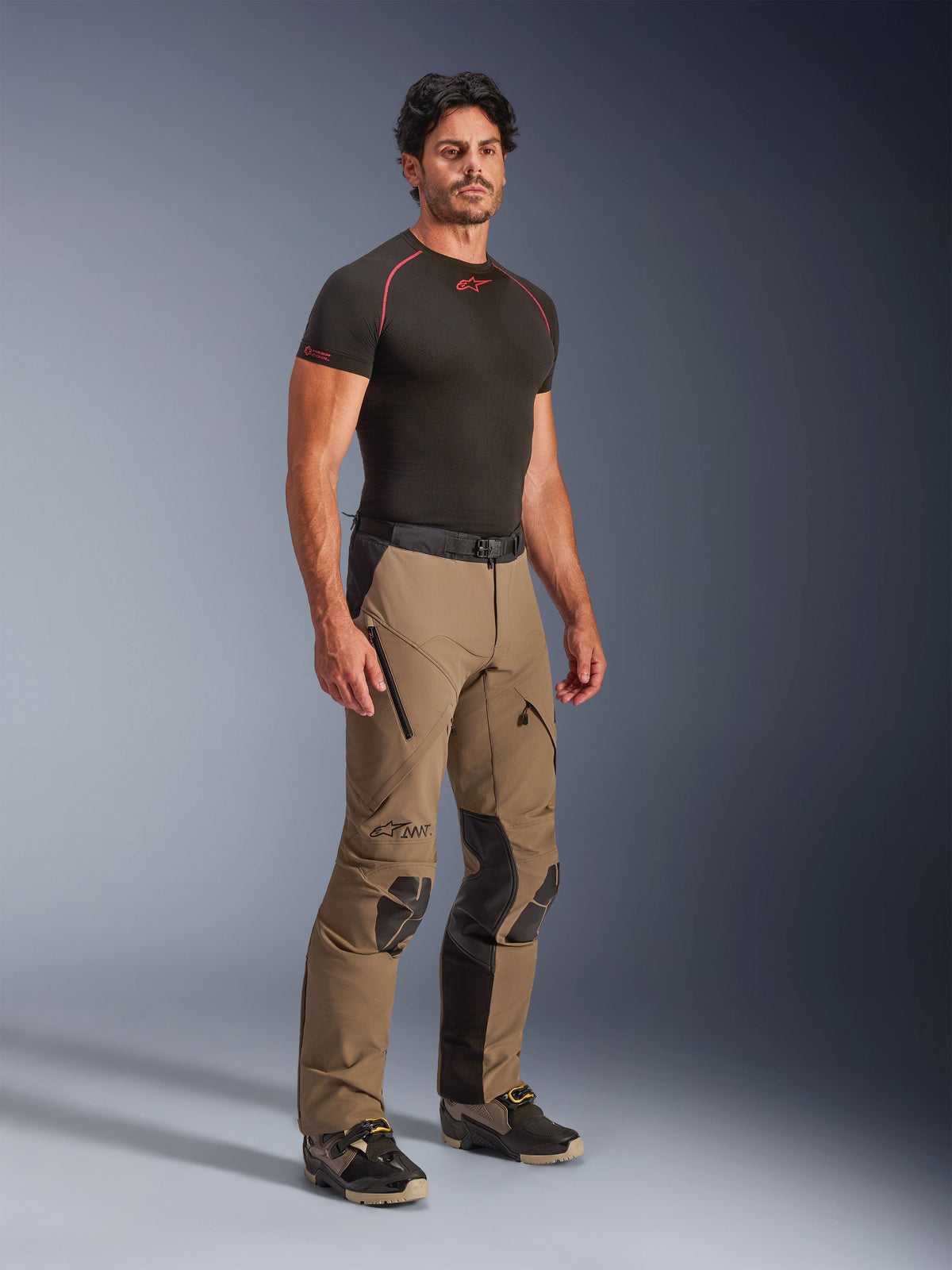Alpinestars AMT-8 Stretch Drystar® XF Hose in Walnut Black, Adventure-Touren-Motorradhose, getragen von einem Model mit einem schwarzen Base-Layer-T-Shirt, mit Stretchmaterial, Belüftungsöffnungen mit Reißverschluss an den Oberschenkeln und schützenden Knieeinsätzen.