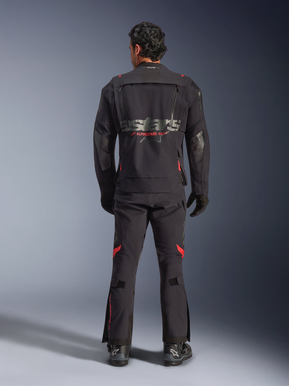 Ein Fahrer in der Rückansicht trägt die Alpinestars Halo Pro Drystar® XF Laminatjacke in Black Dark Shadow, mit einem großen grauen Astars-Logo und rotem Alpinestars-Schriftzug, modularen Belüftungspanelen am Rücken, kombiniert mit einer passenden Adventure-Fahrhose und schwarzen Stiefeln.