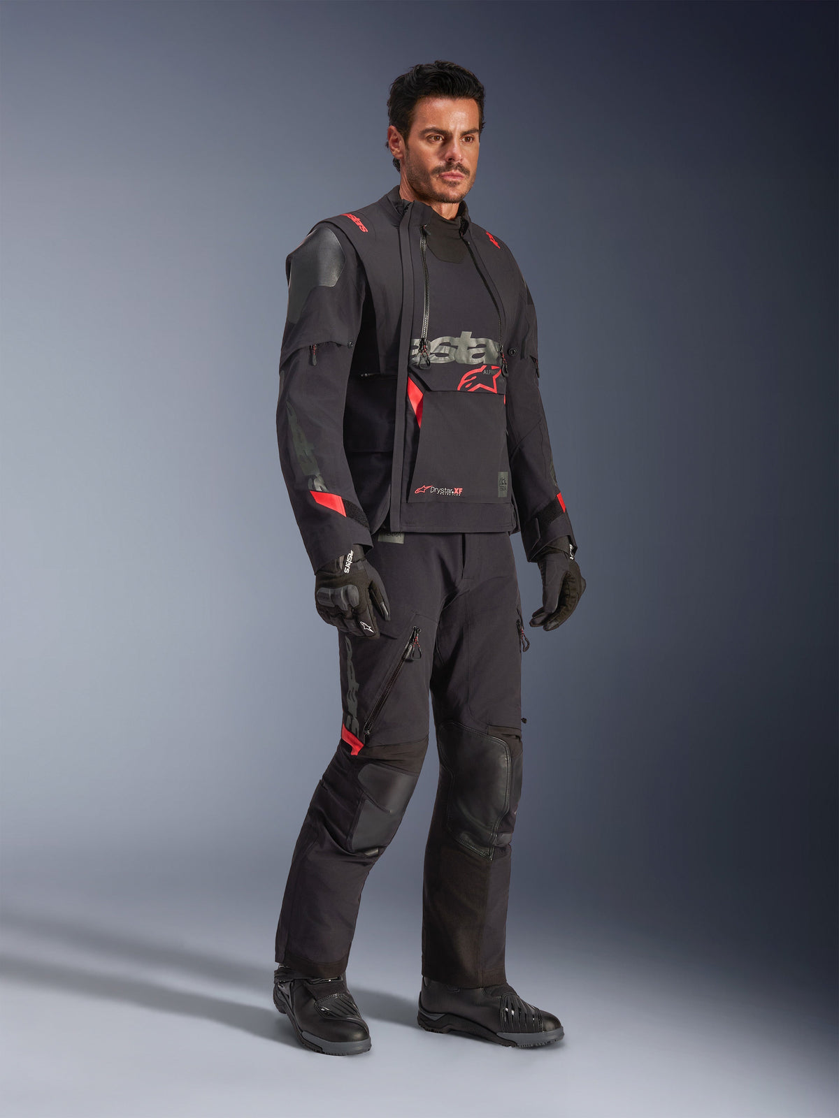 Männlicher Motorradfahrer in der Alpinestars Halo Pro Drystar® XF Laminierten Jacke in Black Dark Shadow, komplette Adventure-Ausrüstung mit passender Hose, Handschuhen und Stiefeln, mit modularen asymmetrischen Frontpaneelen und roten Markenakzenten.