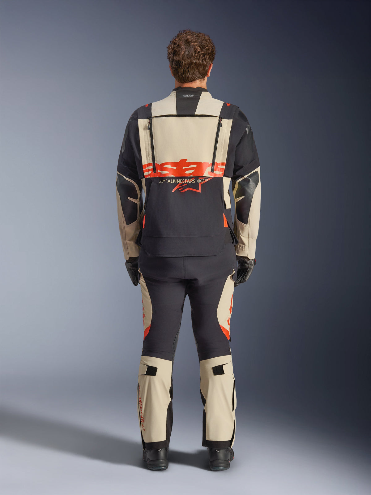 Alpinestars Halo Pro Drystar® XF Laminierte Jacke, Adventure-Motorradjacke, Mastic Black mit orangefarbenem Logo, Rückansicht mit großem Branding am Rücken und modularen Belüftungsreißverschlüssen, getragen mit passender Adventure-Hose für All-Terrain-Performance