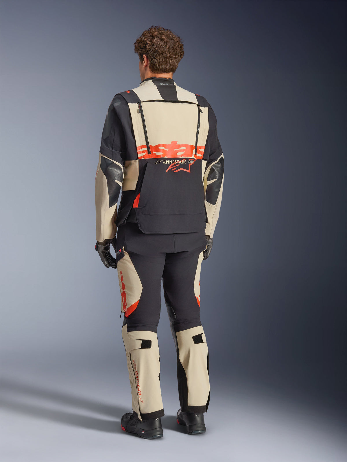 Alpinestars Halo Pro Drystar® XF Laminierte Jacke, Mastic Black, Rückansicht, Adventure-Motorradjacke mit großem orangefarbenem 'astars'-Branding auf der Rückseite, getragen mit passender technischer Hose und Stiefeln, modulares Design für vielseitiges Gelände