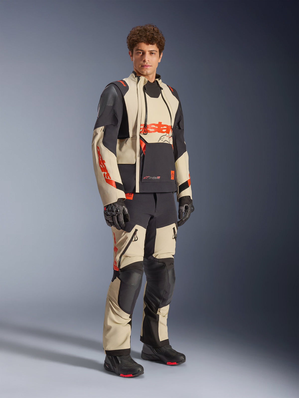 Männliches Model in der Alpinestars Halo Pro Drystar® XF Laminierten Jacke in Mastic Black, mit beigen und schwarzen Einsätzen sowie roten Akzenten, kombiniert mit einer passenden Adventure-Hose und schwarzen Handschuhen, entwickelt für All-Terrain-Abenteuer-Motorradfahrten.
