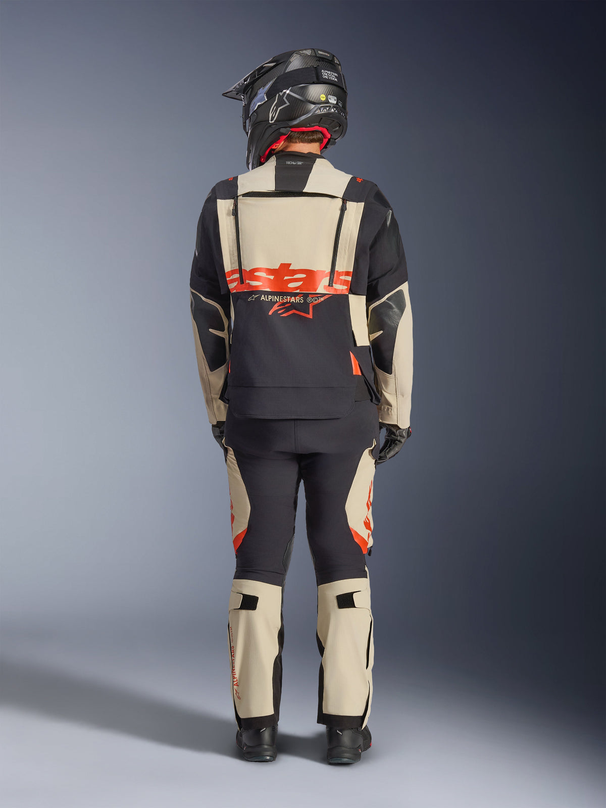 Alpinestars Halo Pro Drystar® XF Laminierte Jacke, Adventure-Motorradjacke, Mastic Black, Rückansicht eines Fahrers, der eine passende Hose und einen schwarzen Helm trägt, mit auffälligem orangefarbenem Alpinestars-Branding und modularen Belüftungspanelen für vielseitige Abenteuerfahrten.