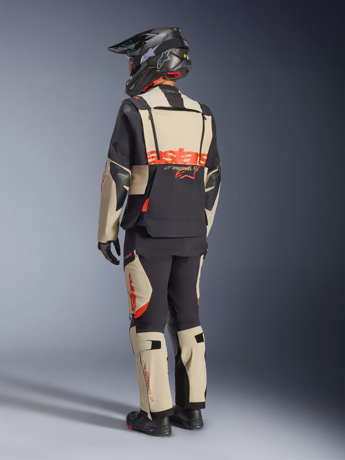 Alpinestars Halo Pro Drystar® XF Laminierte Jacke, Mastic Black, Rückansicht eines Fahrers im kompletten Adventure-Motorradanzug mit passender Hose, schwarzem Adventure-Helm und Stiefeln, mit einem markanten roten Alpinestars-Logo auf dem Rücken.