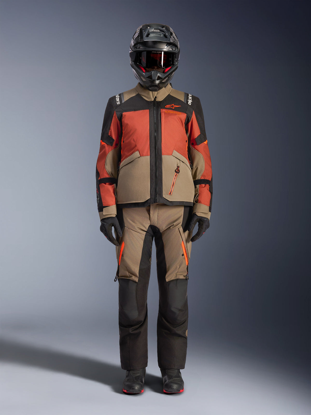 Adventure-Fahrer in der Alpinestars Andes V4 Drystar® Jacke in Walnut Rust Black, kombiniert mit der passenden Adventure-Hose, einem schwarzen Helm mit roten Akzenten und Goggles sowie schwarzen Handschuhen; präsentiert ein komplettes Allwetter-Touren-Outfit.