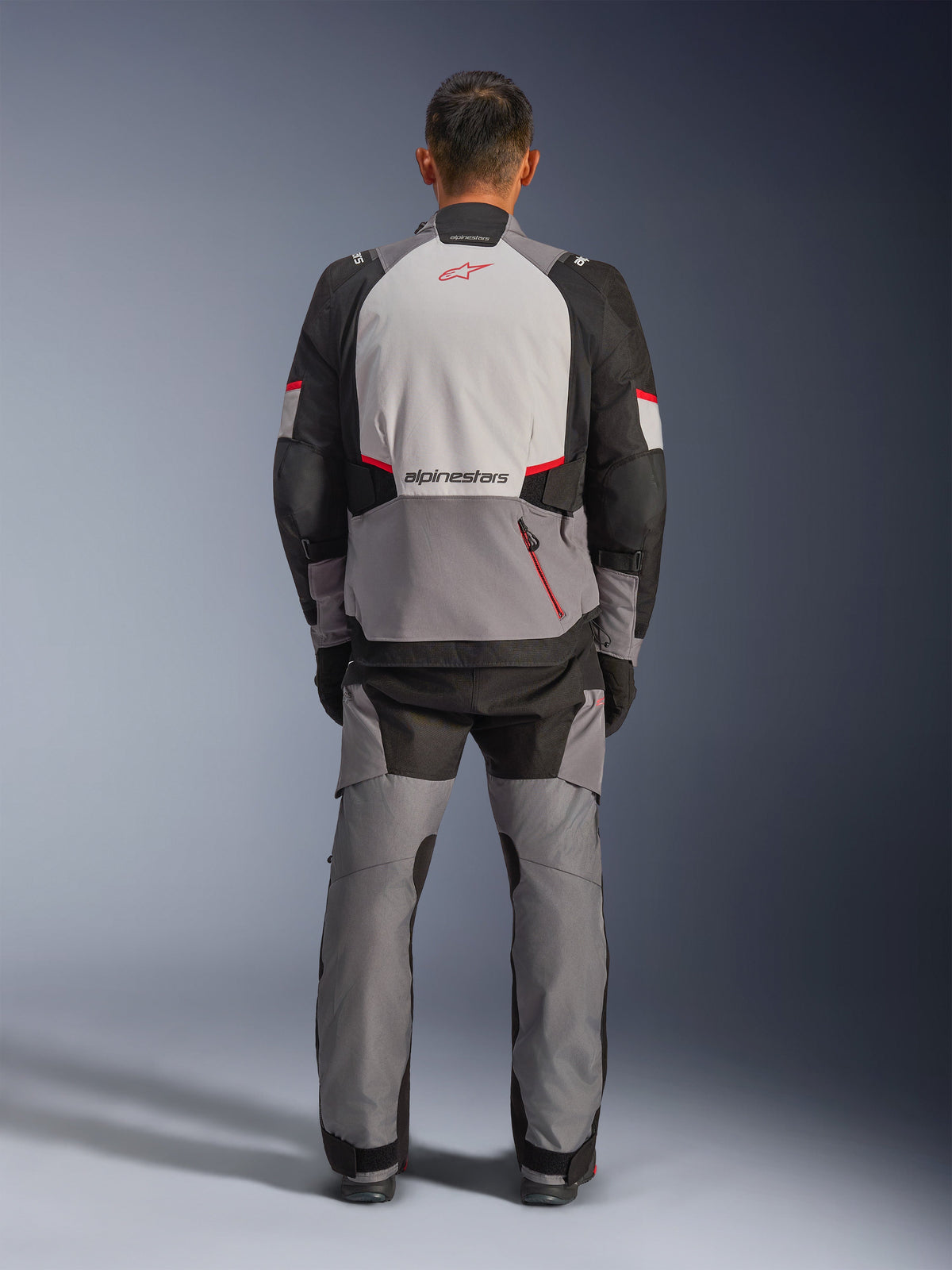 Alpinestars Andes V4 Drystar® Jacket, Adventure Motorcycle Jacket, Dark Gray Ice Gray Black and Bright Red, rear view of a rider wearing matching pants, featuring a large ice gray back panel with red logo, black reinforced shoulder sections, and adjustable waist straps for all-weather touring.
