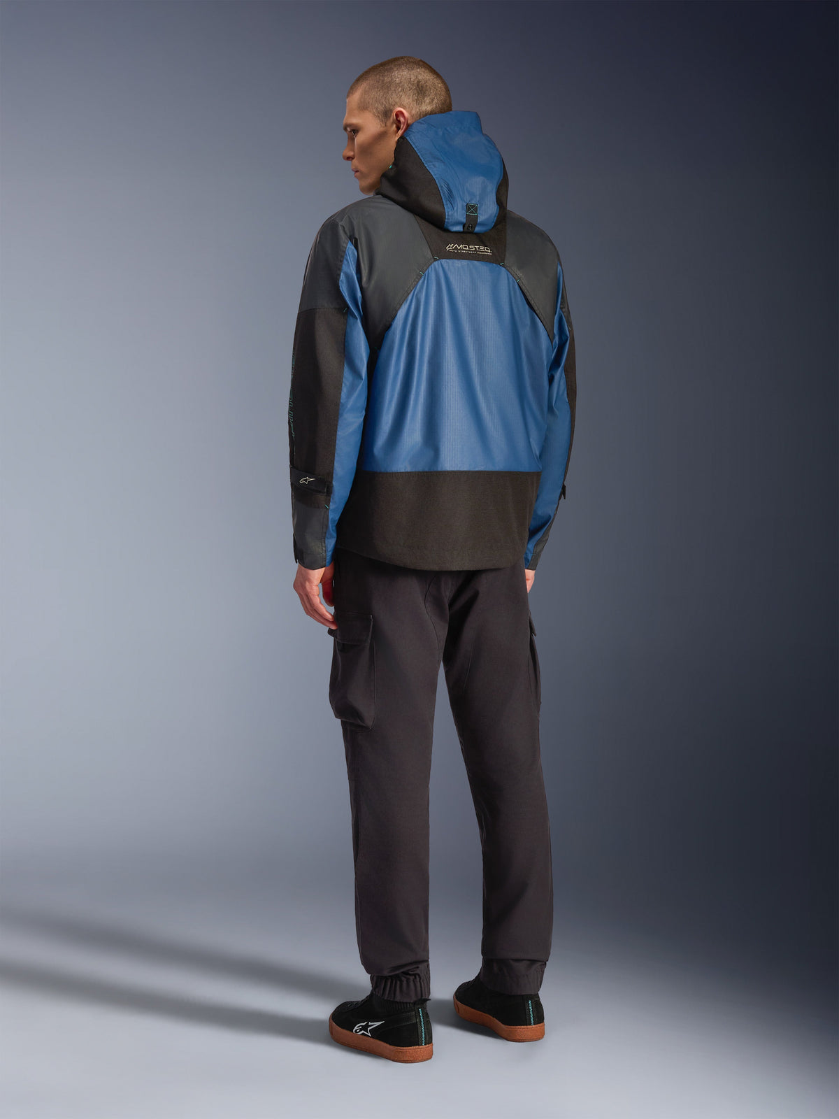 Rückansicht eines männlichen Models in der Alpinestars Mospher Waterproof Jacke in Schwarz und Dark Petrol, mit Kapuzendesign und taktischer Ripstop-Konstruktion, kombiniert mit einer schwarzen Cargohose und Sneakern.