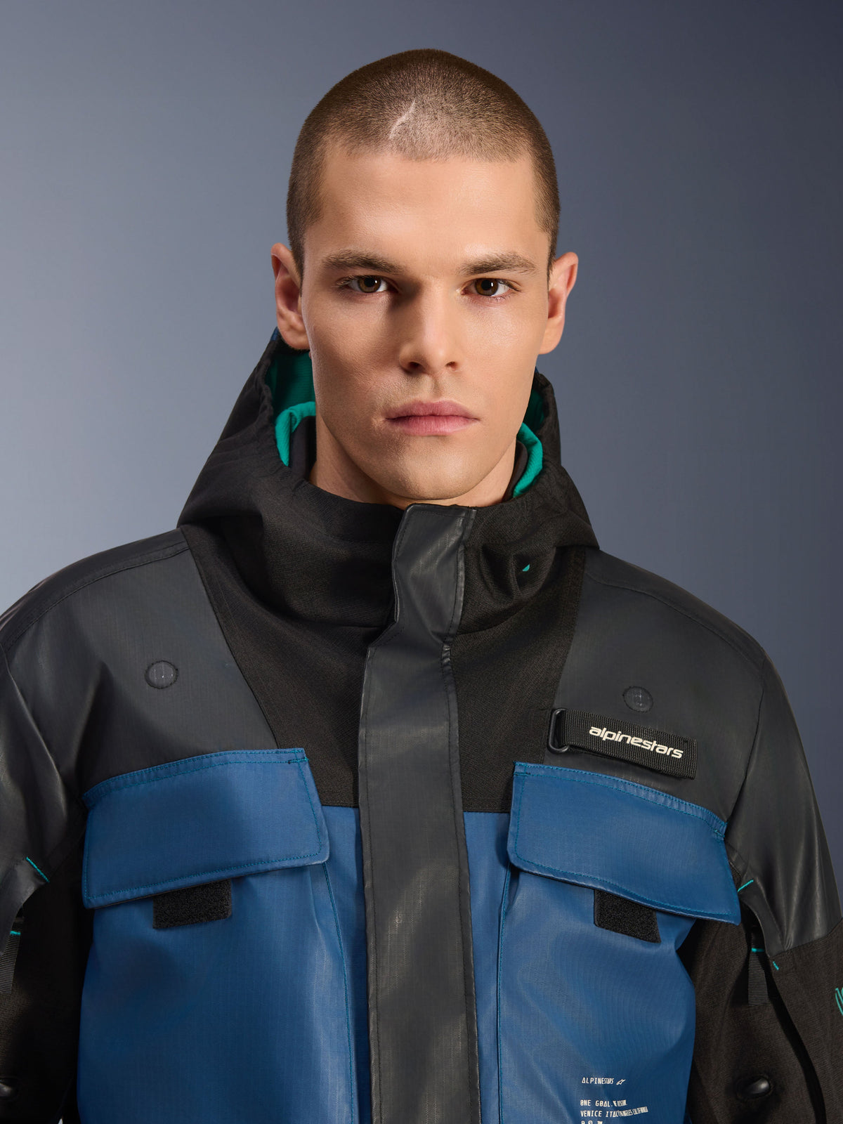 Alpinestars Mospher Waterproof Jacke, Schwarz und Dark Petrol, Nahaufnahme eines männlichen Models in dem Utility-inspirierten Parka mit dunkelblauen Brusttaschen, schwarzer Kapuze mit petrolfarbenem Futter und Alpinestars-Branding auf der Brust.