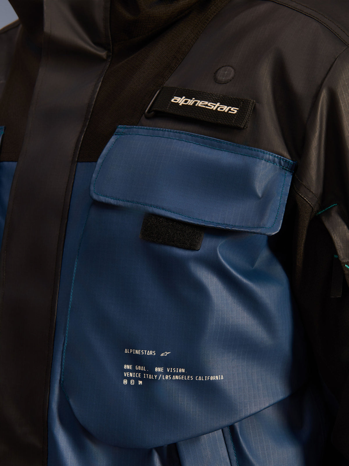 Alpinestars Mospher Waterproof Jacke, Nahaufnahme des Brusttaschenbereichs, Schwarz und Dark Petrol, strapazierfähiges Ripstop-Gewebe mit aufgedrucktem „One Goal. One Vision.“ Schriftzug, nutzungsorientiertes Design mit funktionellen Taschen und Alpinestars Logo-Lasche
