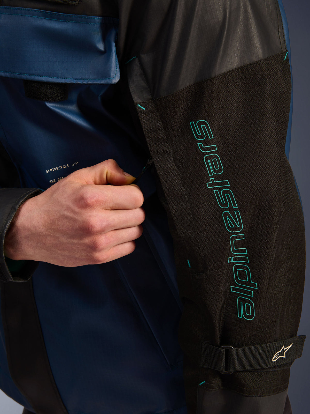 Alpinestars Mospher Waterproof Jacke in Black Dark Petrol, Detailaufnahme des Ärmels mit türkisem Alpinestars-Branding, eine Hand verstellt den Belüftungsreißverschluss, strapazierfähiges Ripstop-Gewebe und verstellbarer Ärmelriemen