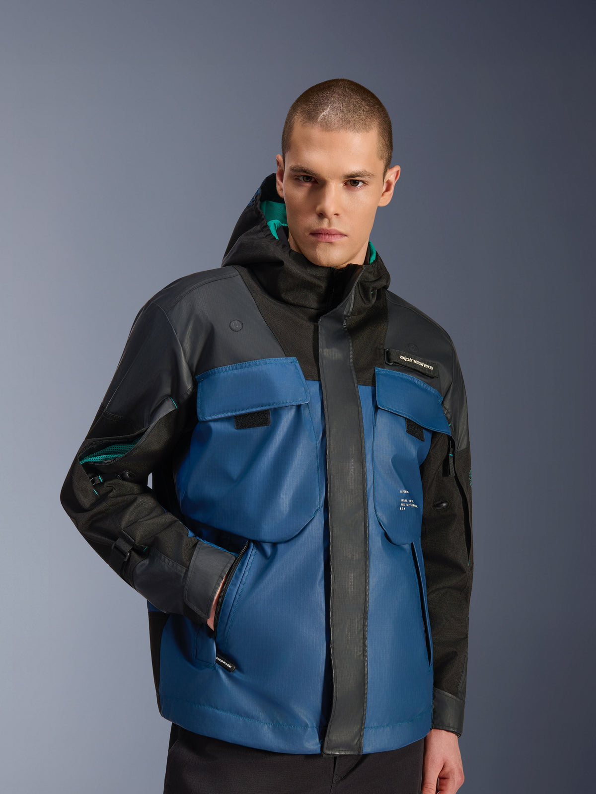 Männliches Model in der Alpinestars Mospher Waterproof Jacke in Schwarz und Dark Petrol, mit großen Utility-Brusttaschen, einem Kapuzendesign mit petrolfarbenem Futter und technischem Ripstop-Gewebe für das urbane Pendeln mit dem Motorrad.