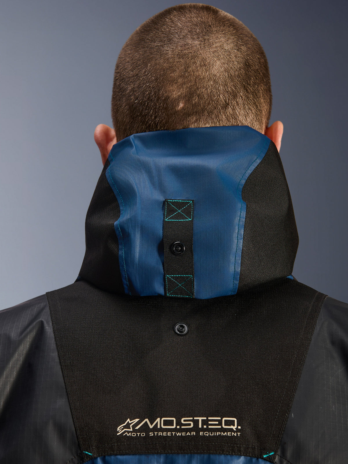 Alpinestars Mospher Waterproof Jacket, Detailansicht der Rückseite mit Kapuzenverankerungssystem, Schwarz und Dark Petrol Blau, MO.ST.EQ. Moto Streetwear Equipment Branding, strapazierfähiges Ripstop-Textilmaterial, technische Motorradbekleidung für Pendler