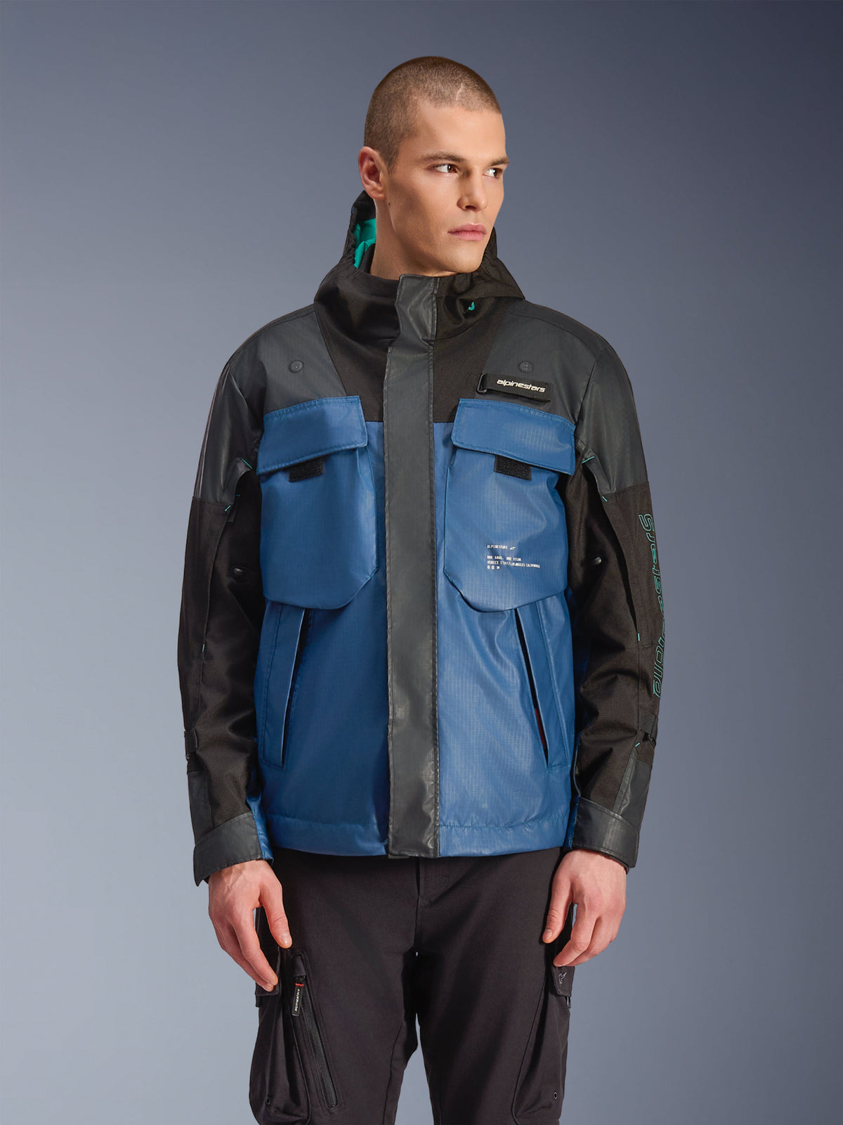 Alpinestars Mospher Waterproof Jacke, Urbaner Motorrad-Parka, Schwarz und Dunkles Petrolblau, mit großen taktischen Brusttaschen, hohem Kragen mit Kapuze, technischer Ripstop-Struktur, Vorderansicht am Modell