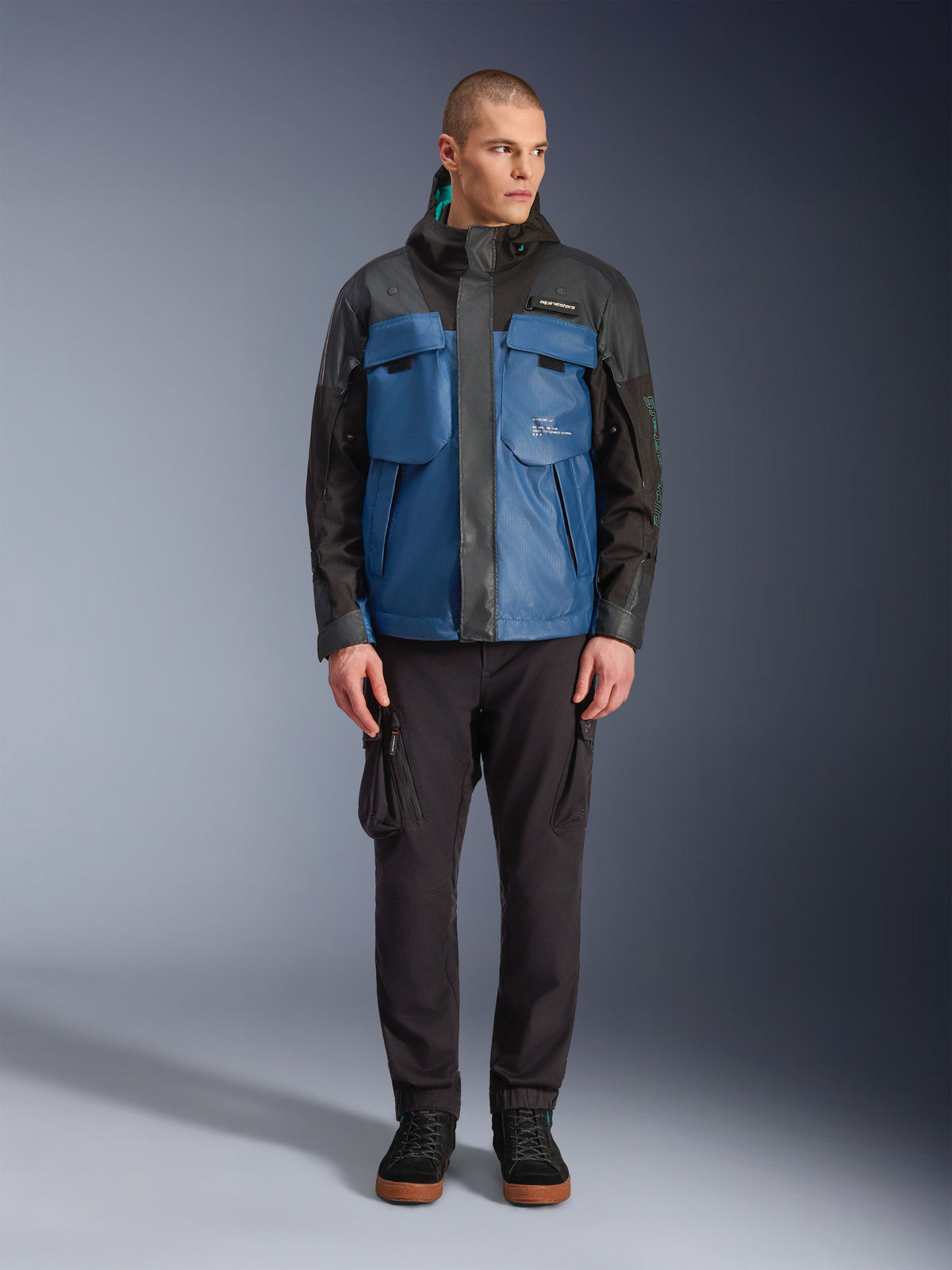 Alpinestars Mospher Waterproof Jacke in Black Dark Petrol, männliches Model trägt einen taktisch inspirierten Ganzjahres-Motorradparka mit großen funktionellen Taschen und integrierter Kapuze, kombiniert mit einer schwarzen technischen Hose und schwarzem Schuhwerk, Studio-Frontansicht