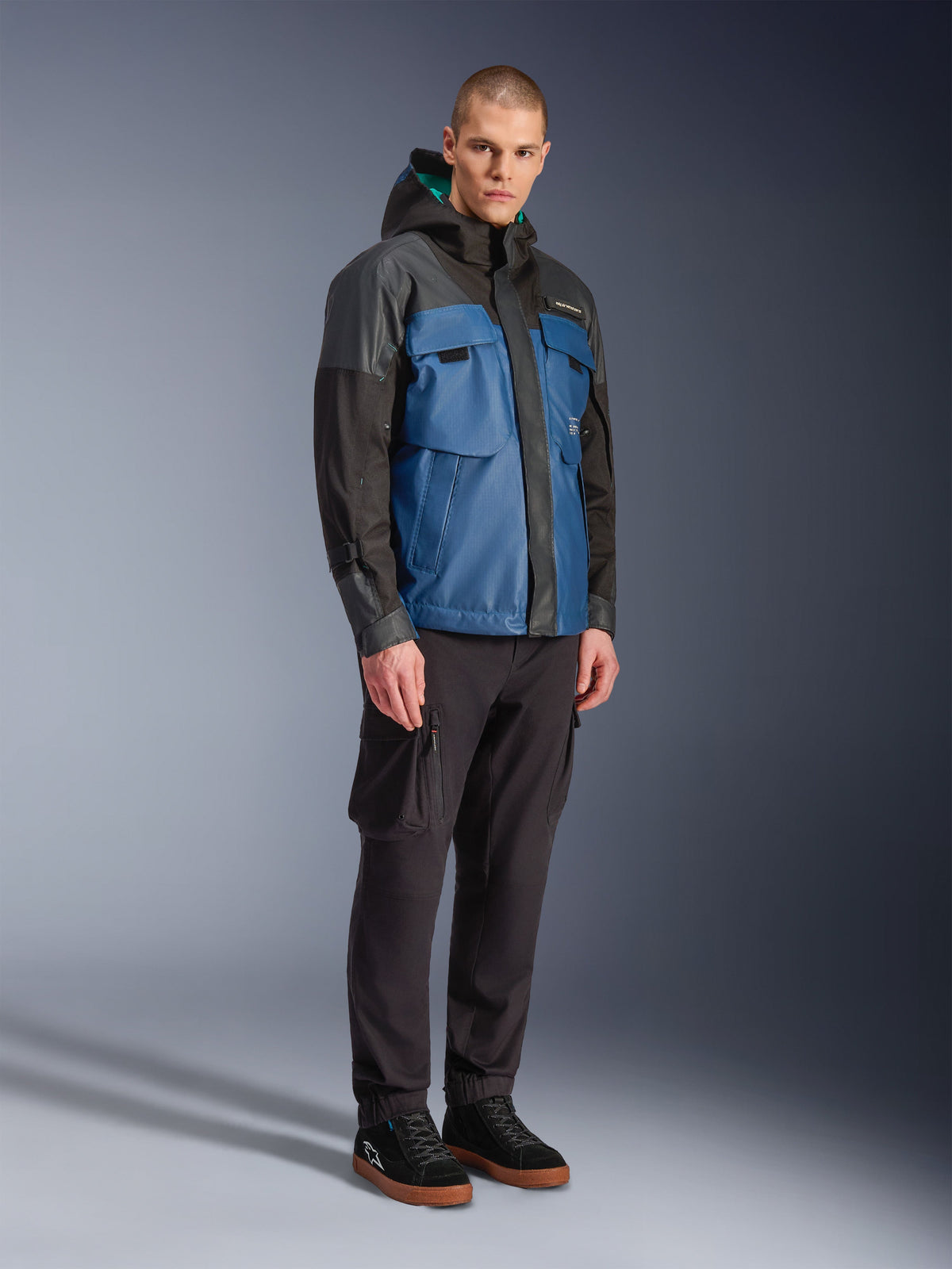 Alpinestars Mospher wasserdichte Jacke, Schwarz und Dunkles Petrolblau, kombiniert mit schwarzer Cargohose und schwarzen Sneakern, urbaner Pendler-Stil mit Funktionstaschen und Kapuze