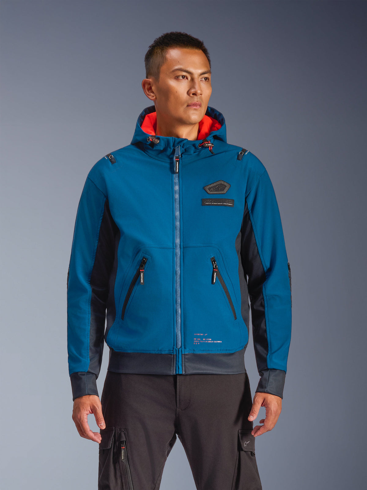 Alpinestars Moblast Waterproof Jacke an einem Model, Blau und Dunkelnavy, Urban-Motorradjacke, Kapuzendesign mit rotem Innenfutter, schwarze Reißverschlüsse und Brust-Patch-Detail, Streetwear-inspirierte Motorradausrüstung
