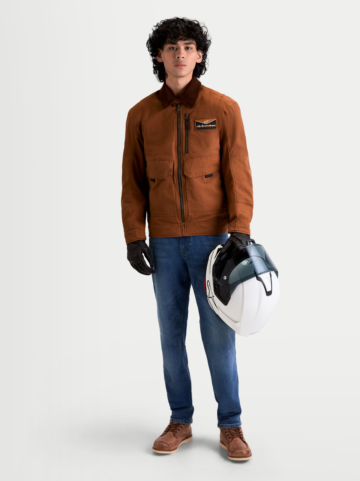 Alpinestars Mustang Wp Jacke, urbane urbane Textil-Motorradjacke für Herren in Rostbraun und Dunkelbraun, Vorderansicht eines Models mit Arbeitskleidung-gestylter Schutzausrüstung, das einen weißen Helm hält