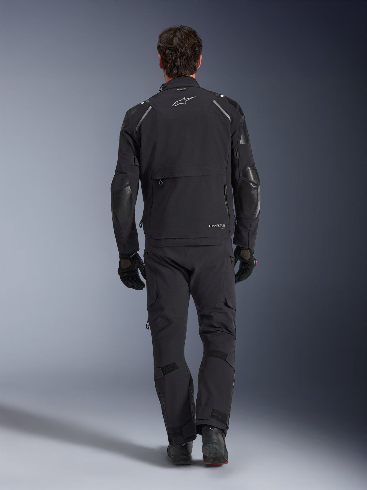 Alpinestars Andes Pro Drystar® XF Jacke, Adventure-Tourenjacke, Schwarz und Dunkelgrau, Rückansicht mit großer Cargotasche und verstärkten Ellbogen-Einsätzen, getragen mit passender schwarzer Hose und Handschuhen, elegantes funktionelles Design für Fahrten bei jedem Wetter