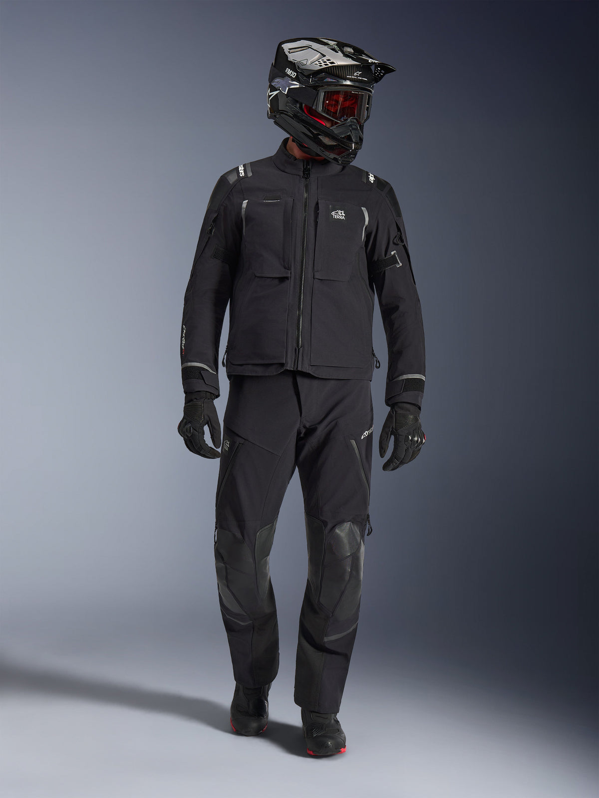 Adventure-Fahrer in der Alpinestars Andes Pro Drystar® XF Jacke in Schwarz und Dunkelgrau, passende schwarze Adventure-Hose, schwarz-weißer Helm mit roter Brille, schwarze Handschuhe und Stiefel, mit großen vorderen Funktionstaschen, reflektierenden Akzenten und technischer 3-Lagen-Laminat-Konstruktion für Allwetter-Touren.
