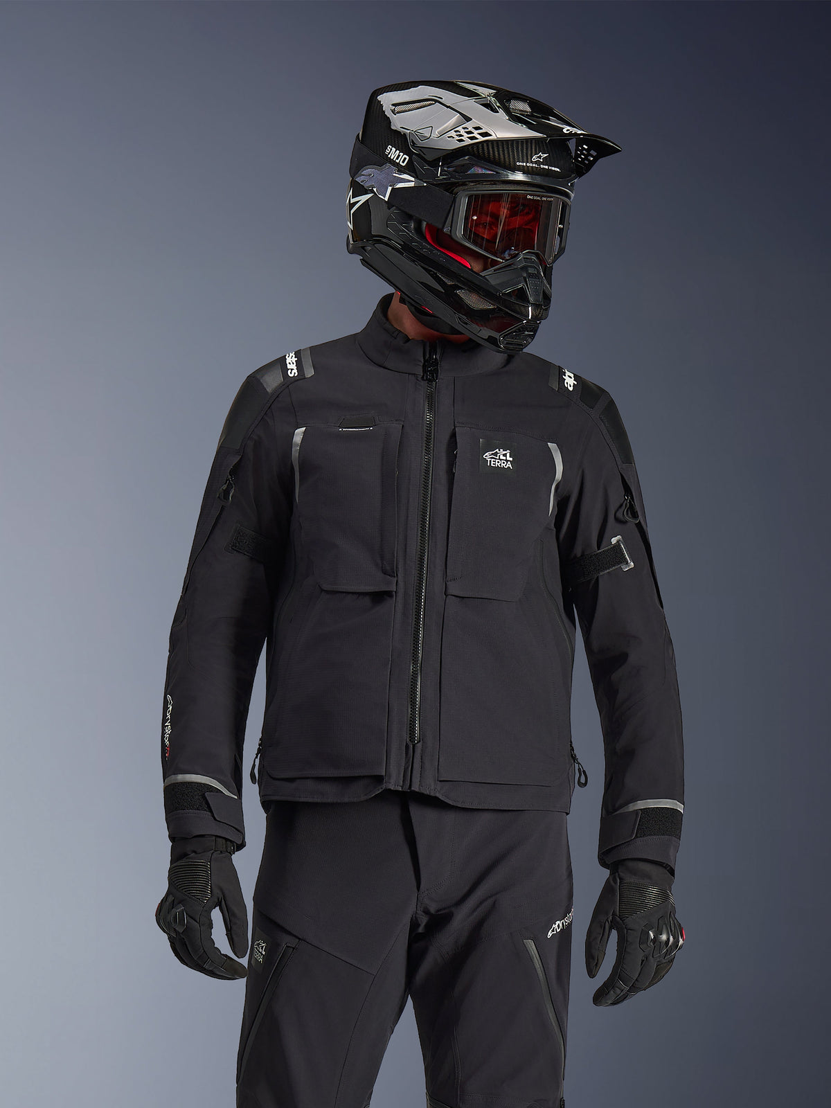 Adventure-Fahrer mit der Alpinestars Andes Pro Drystar® XF Jacke in Schwarz und Dunkelgrau, All-Terra-Branding auf der Brust, kombiniert mit passender technischer Hose und schwarzem Adventure-Helm mit Schutzbrille, professionelles Touren-Outfit.