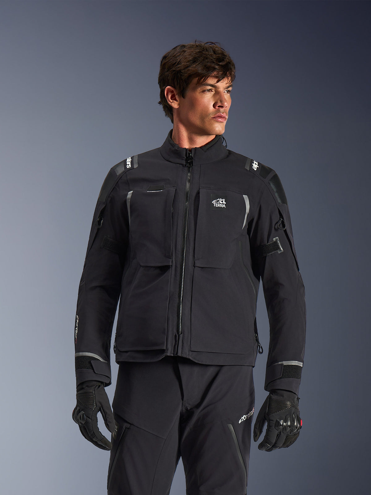 Alpinestars Andes Pro Drystar® XF Jacke, schwarz-dunkelgraue Motorrad-Adventure-Touringjacke, getragen mit passender Hose und Handschuhen, technische laminierte Schalenkonstruktion, ALL TERRA Logo auf der Brust, reflektierende Details, verstellbare Ärmelbündchen