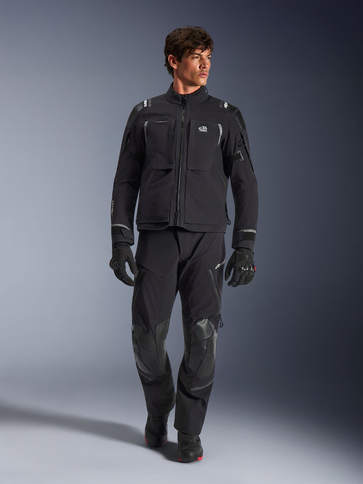 Männliches Model in Alpinestars Andes Pro Drystar® XF Jacke, Adventure-Motorradjacke in Schwarz und Dunkelgrau, kombiniert mit passender technischer Hose und Handschuhen, mit mehreren Taschen und „Terra“-Branding auf der Brust.