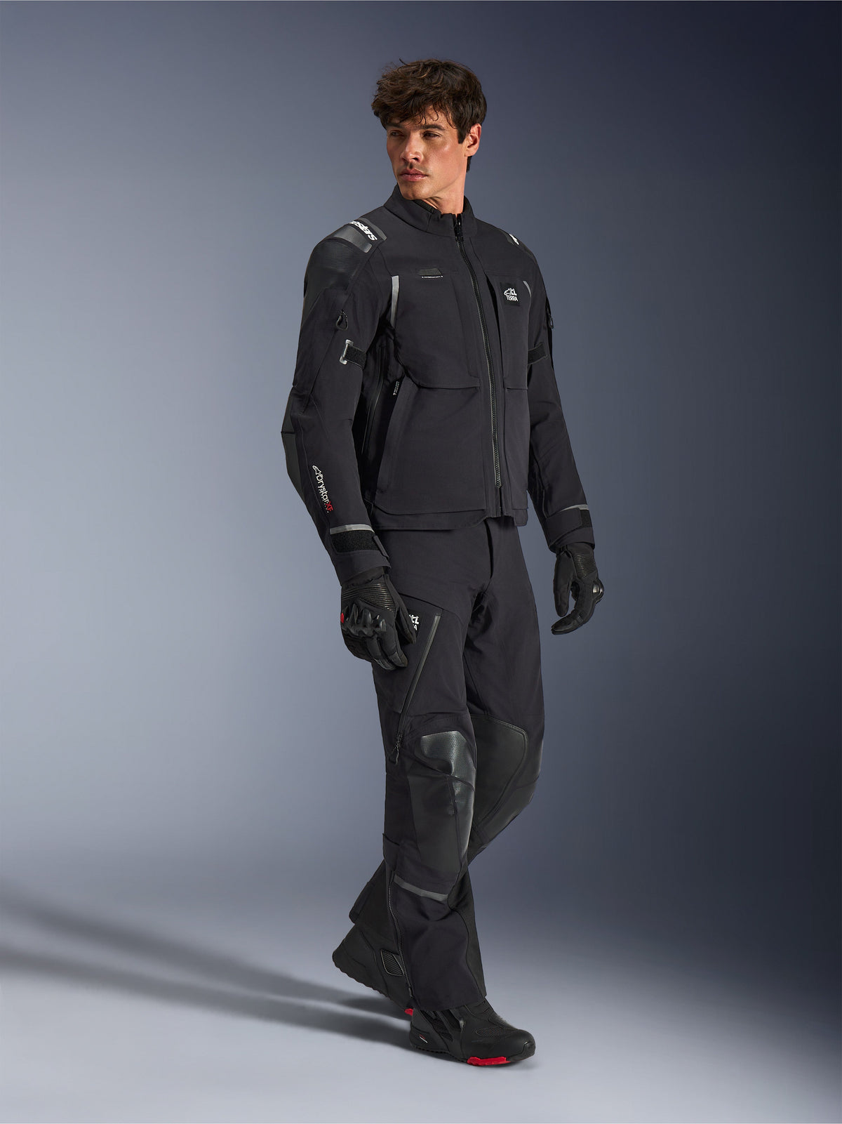 Alpinestars Andes Pro Drystar® XF Jacke, Motorradjacke in Schwarz und Dunkelgrau, getragen von männlichem Model mit passender Adventure-Hose, Handschuhen und Stiefeln, Ganzkörper-Studioaufnahme, technische Adventure-Touring-Ausrüstung mit reflektierenden Details und Branding