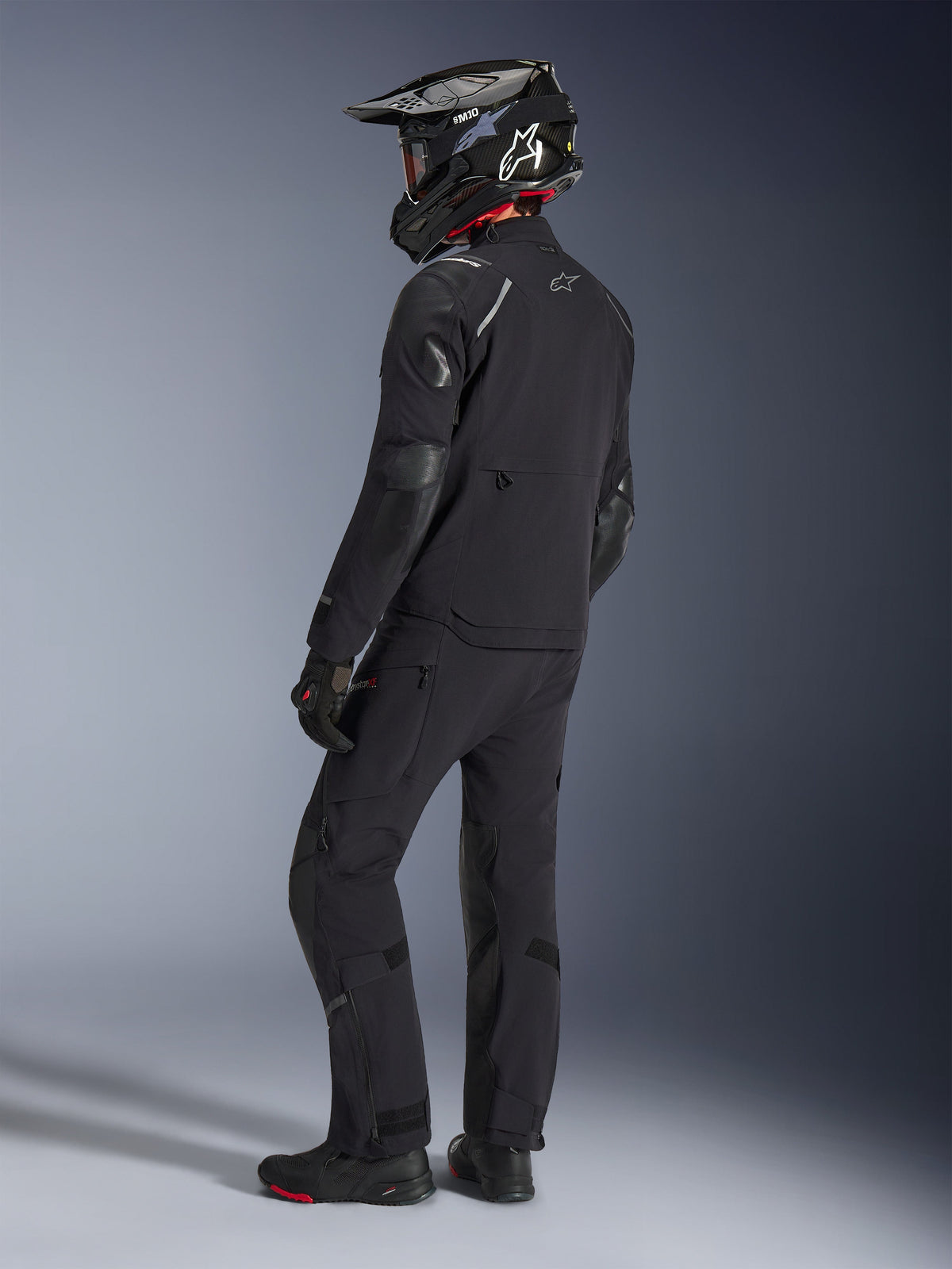 Motorradfahrer in der Alpinestars Andes Pro Drystar® XF Jacke und passender Hose in Schwarz und Dunkelgrau, Rückansicht im Profil, mit schwarzem Carbon-Adventure-Helm, schwarzen Handschuhen und Adventure-Stiefeln, hochwertige abgestimmte Ausrüstung für Adventure-Touren.