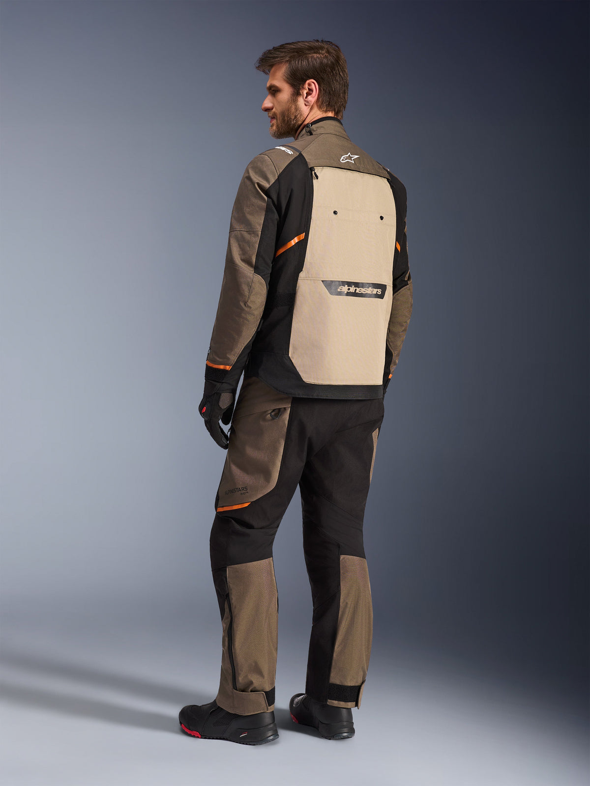Alpinestars Cusco Drystar® Jacke in Walnut Aluminum Black, Rückansicht mit beigem Rückenpaneel und orangefarbenen Akzenten, getragen mit passender Motorradhose, schwarzen Handschuhen und Stiefeln, professionelle Adventure-Touring-Ausrüstung